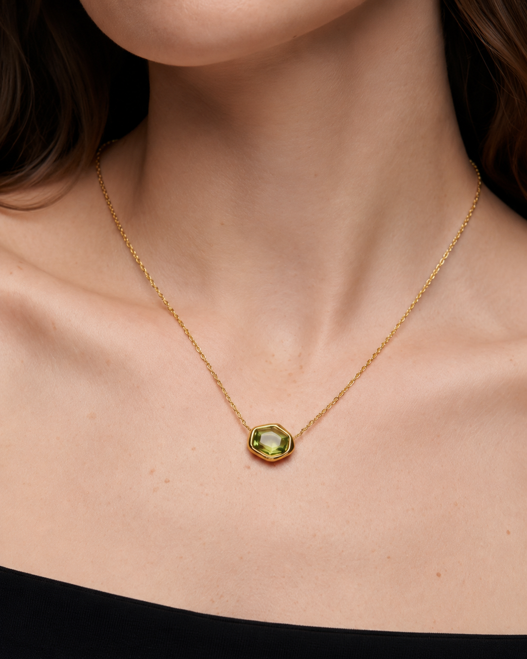 A close-up of a model wearing a delicate gold chain necklace with a faceted olive-green gemstone pendant.