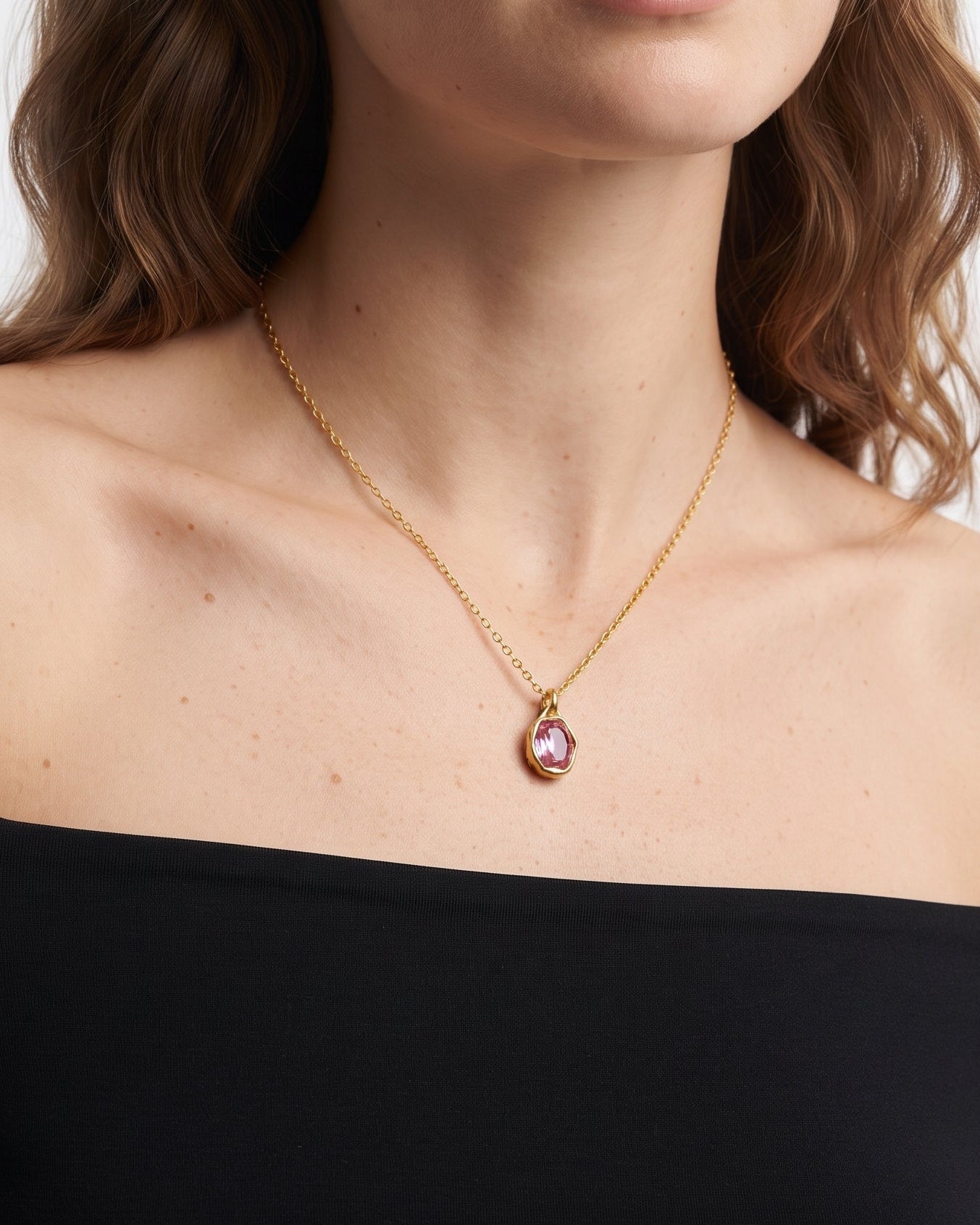 A woman wearing a delicate gold chain necklace with a teardrop-shaped pink gemstone pendant, styled with a black off-the-shoulder top against a light gray background.