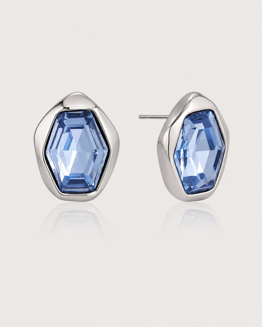 A pair of silver stud earrings with irregular, organic-shaped bezel settings holding faceted light blue gemstones, shown on a light beige background with a soft reflection.