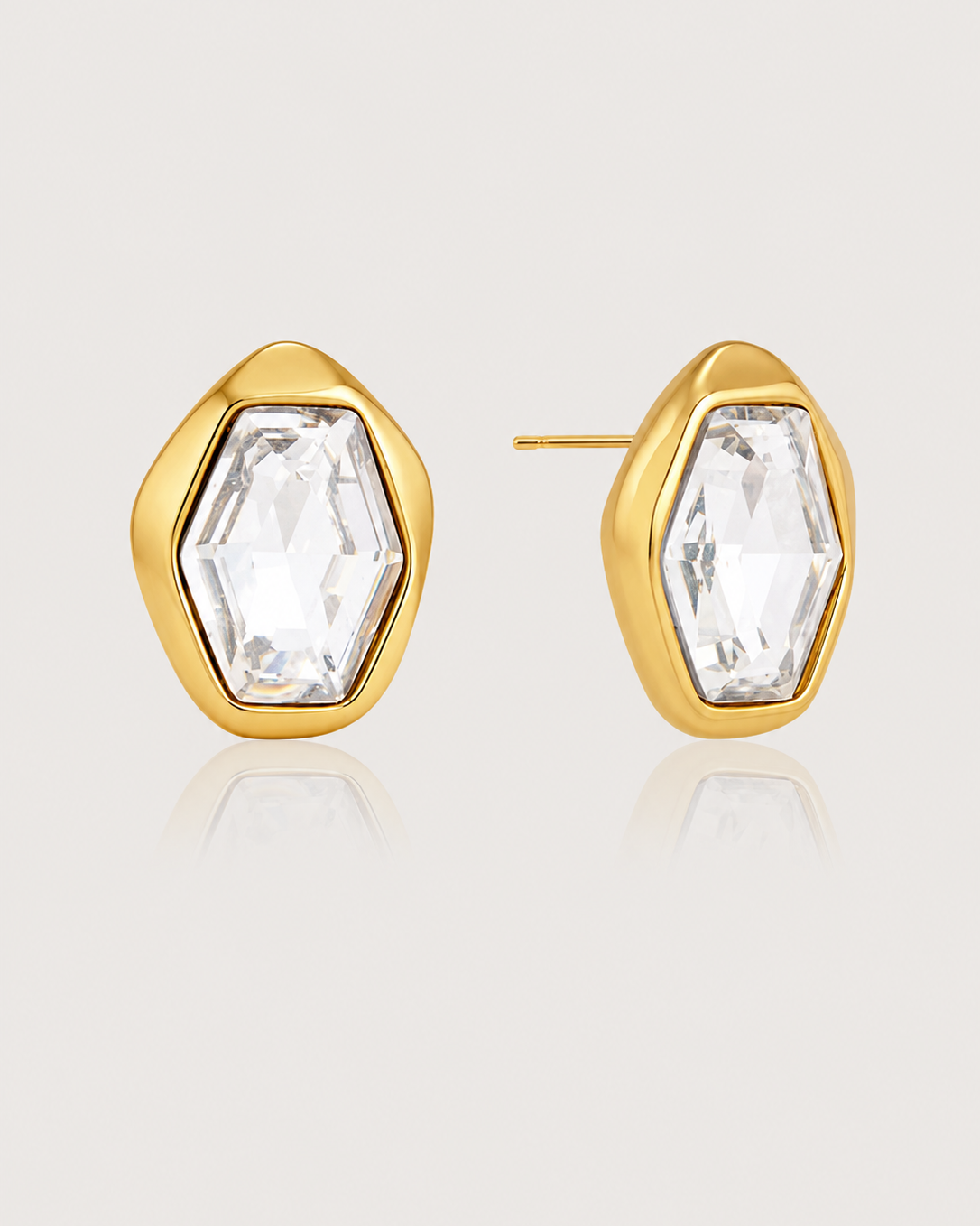 A pair of gold stud earrings with a faceted, clear marquise-cut gemstone in an organic, irregular bezel setting, shown on a light beige background with a soft reflection.