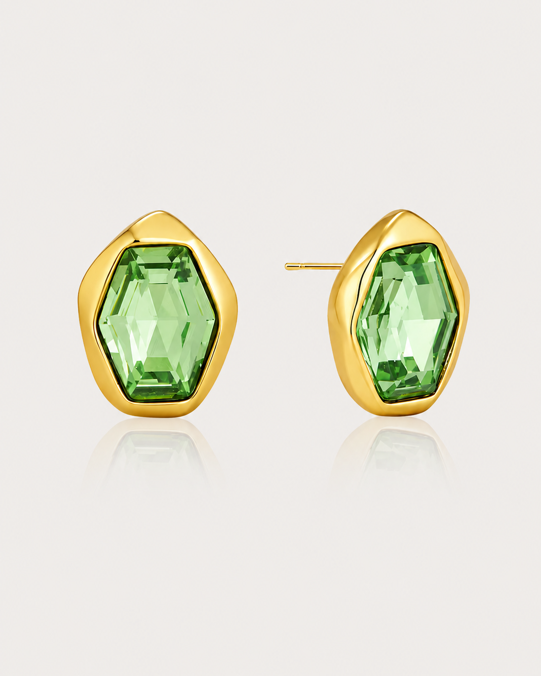 A pair of gold stud earrings with faceted green gemstones in an organic, rounded geometric shape, shown on a light beige background with a soft reflection.