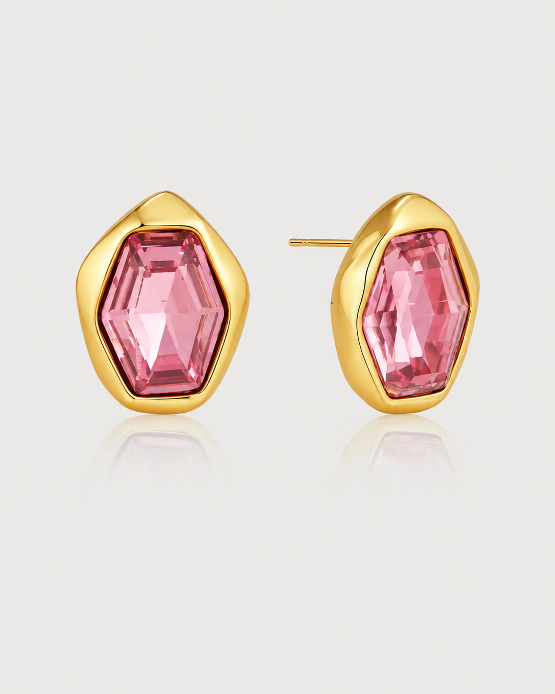 A pair of gold-toned stud earrings with faceted pink gemstones, shown on a light gray reflective background.