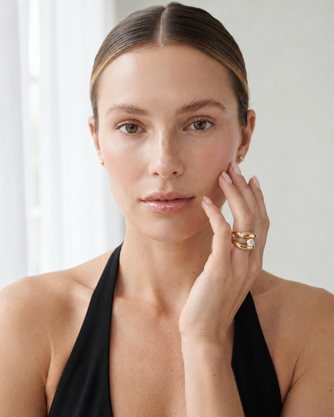 A woman with her hair pulled back wears a delicate gold ring with a clear central stone on her finger, posing in a black halter top against a bright, softly lit background.