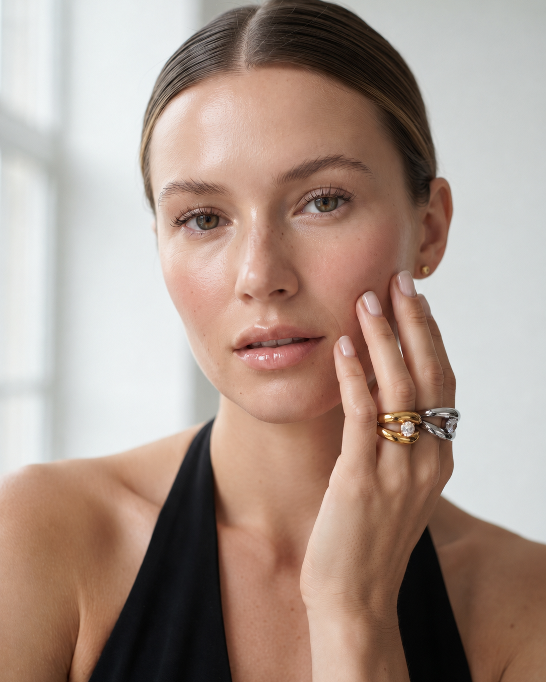 A close-up portrait of a woman wearing two interlocking rings on her finger. One ring is gold with a clear stone, and the other is silver with a clear stone.
