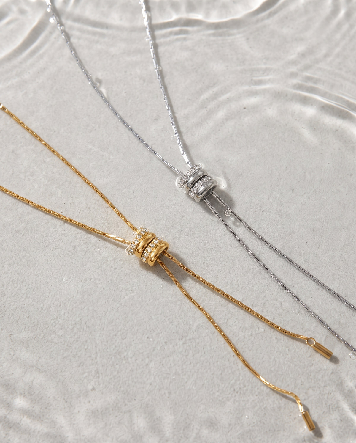 Two delicate chain necklaces with wrapped cylindrical links and drop chains, one in gold and one in silver, laid on a light textured surface with subtle water ripples.