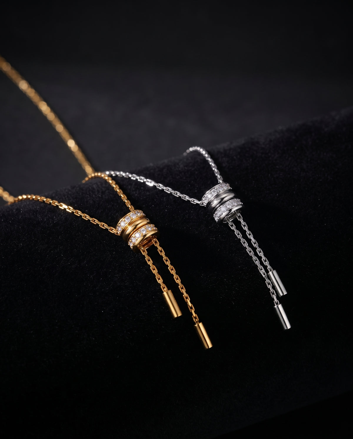Two drop chain necklaces displayed on a black velvet surface, one in gold and one in silver, both featuring stacked cylindrical links with small clear stones and tapered drop chains.
