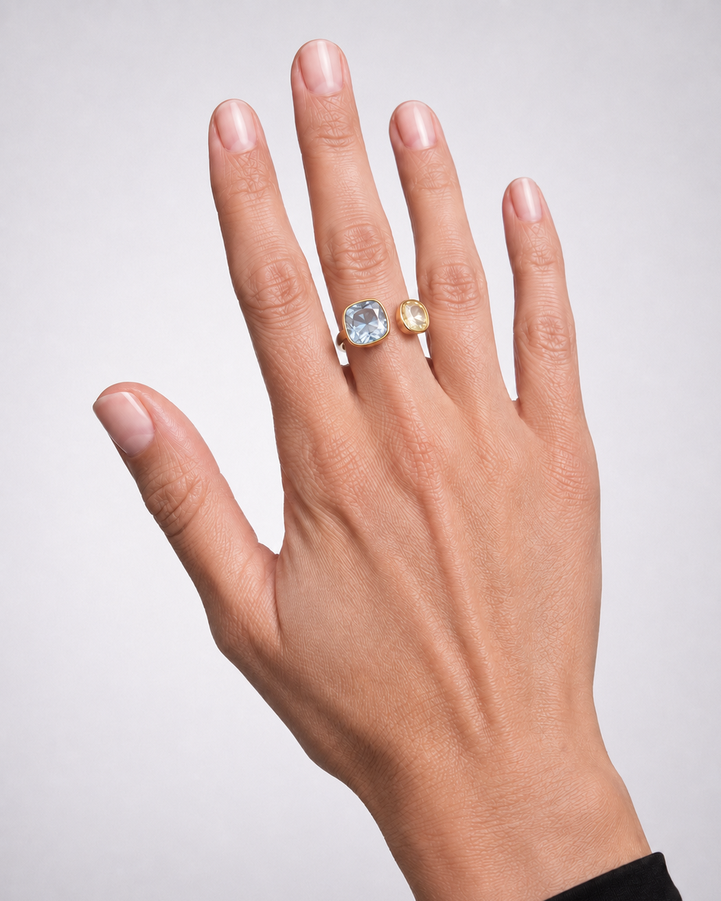 A close-up of a hand wearing two rings: a larger silver-toned ring with a round clear gemstone and a smaller yellow gold ring with a round clear gemstone, set against a light gray background.