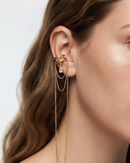 A close-up of a model wearing a gold ear cuff with multiple draped chain earrings, set against a clean light gray background.