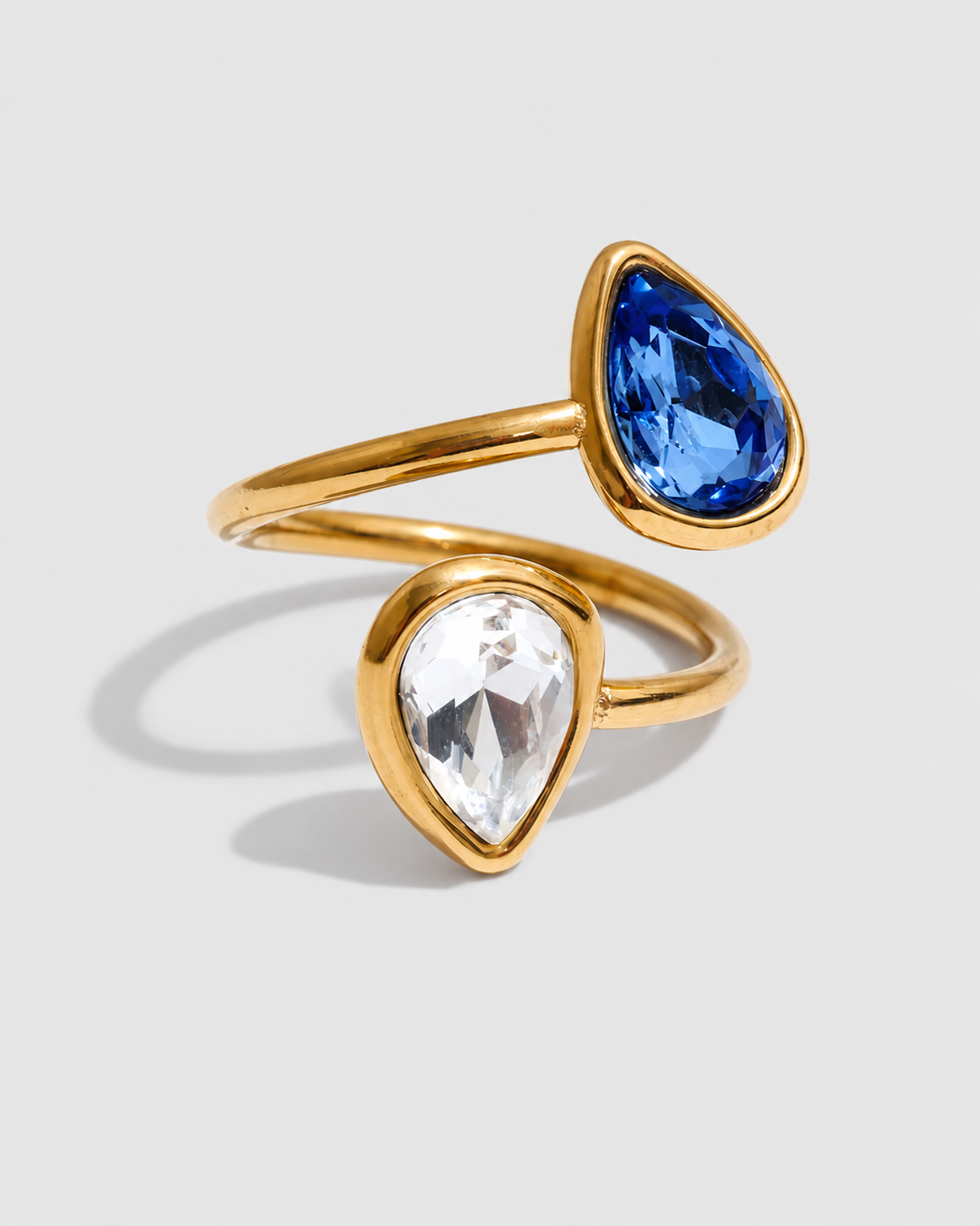 A gold-plated double-stone ring featuring one pear-cut blue gemstone and one pear-cut clear gemstone, set on a light gray background.
