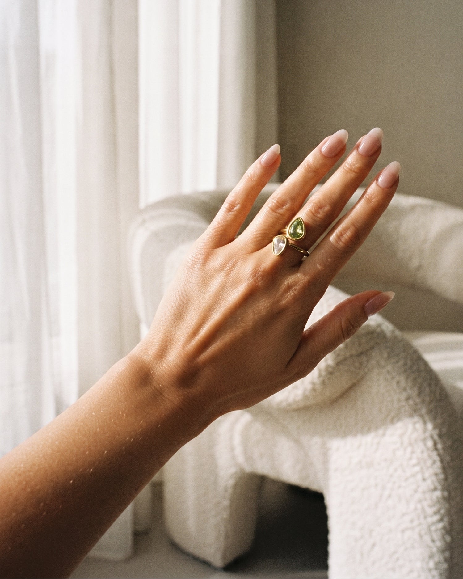A close-up of a person's hand wearing two rings, one with a green marquise-cut stone and one with a small clear stone, set against a softly lit room with a textured white chair and sheer curtains.