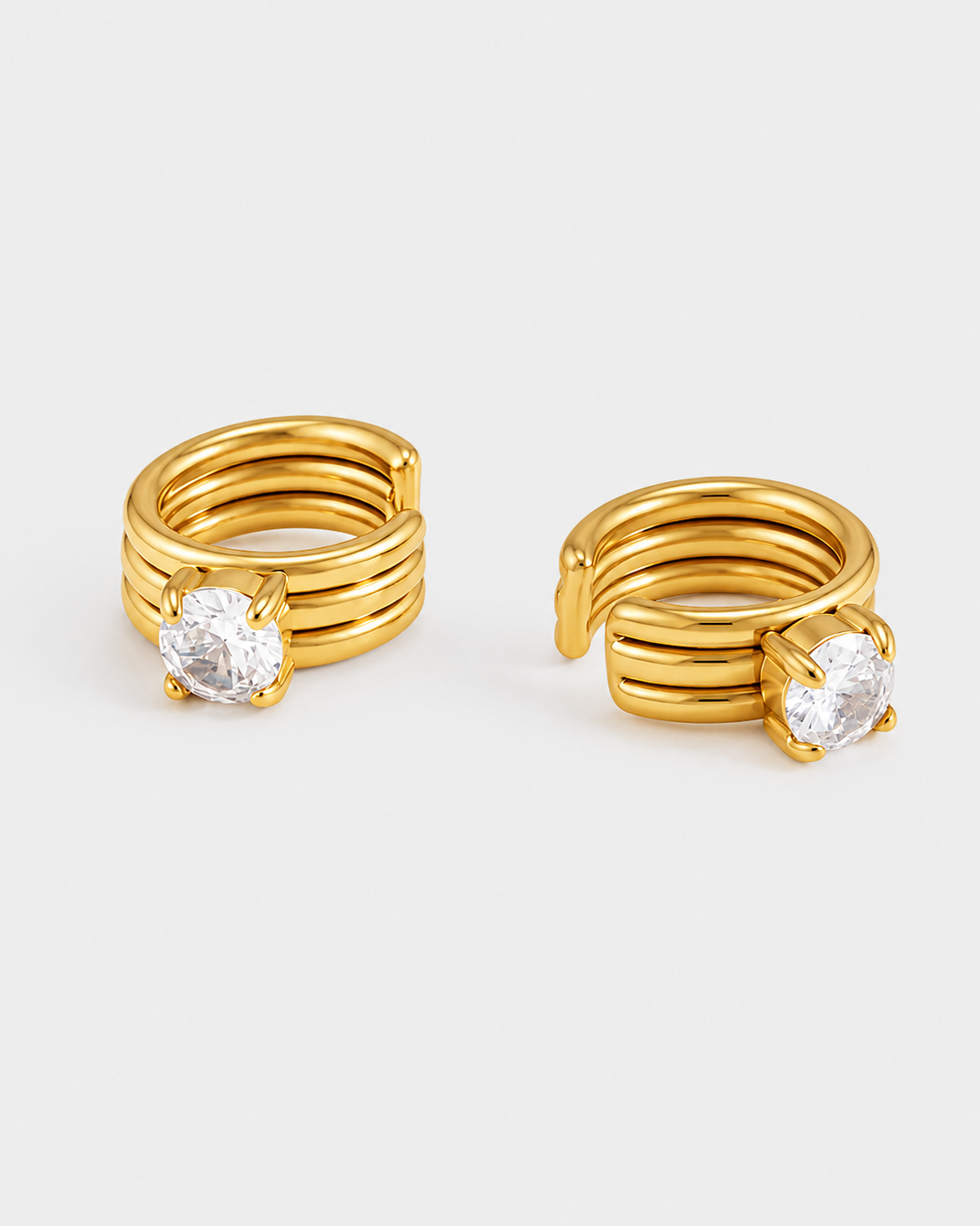 A pair of gold huggie hoop earrings with a multi-band wrap design and a clear round-cut gemstone on each earring, shown on a light gray background.