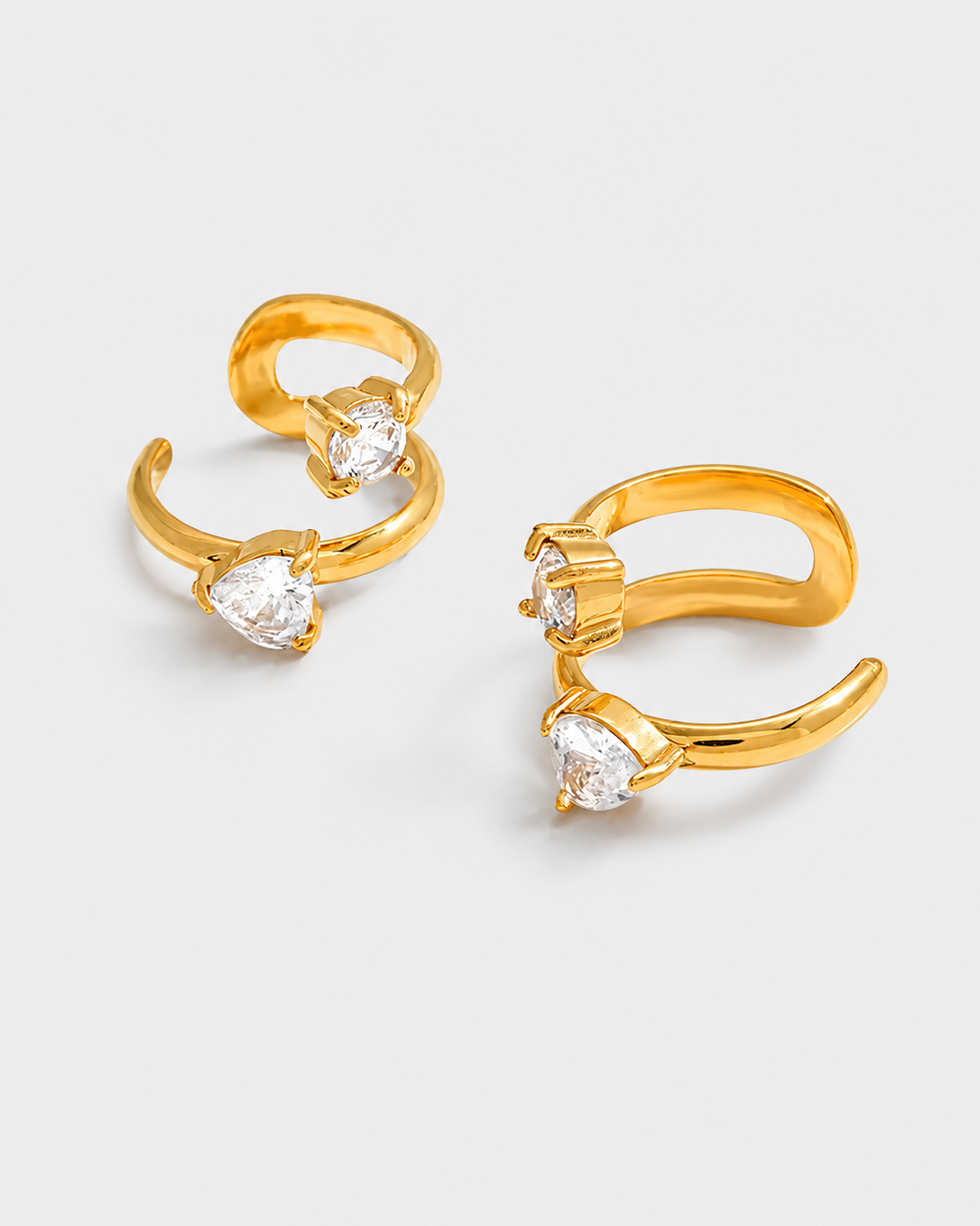 A pair of gold huggie hoop earrings, each featuring a curved gold band and a clear heart-shaped gemstone at the tip, set against a light gray background.
