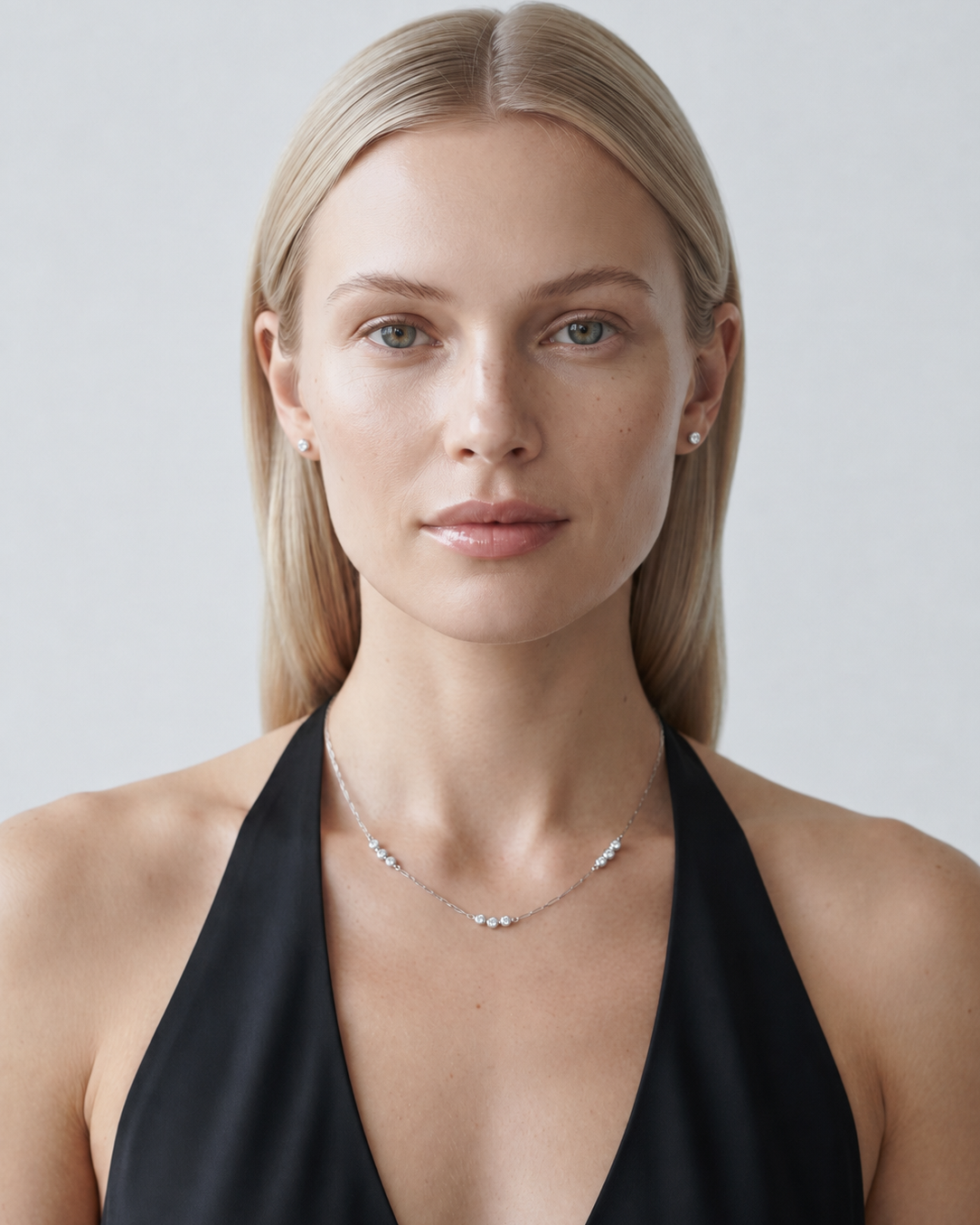 A blonde woman wearing a delicate silver necklace with small clear stones and a black halter top, photographed against a light gray studio background.
