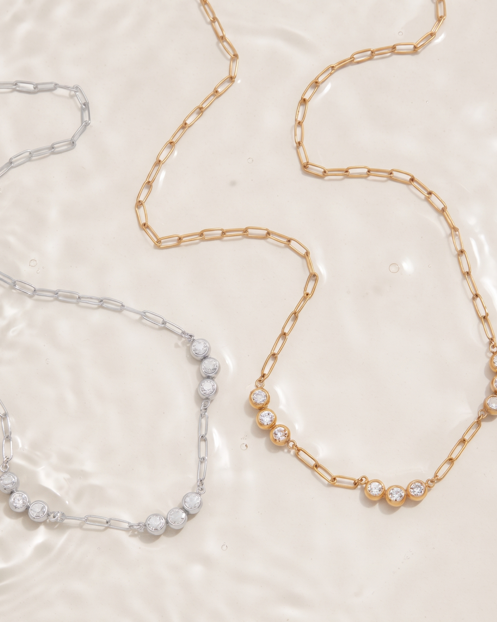 Two delicate chain necklaces, one gold and one silver, laid out on a glossy, reflective cream surface. Each necklace features a section of small, polished round beads at one end.