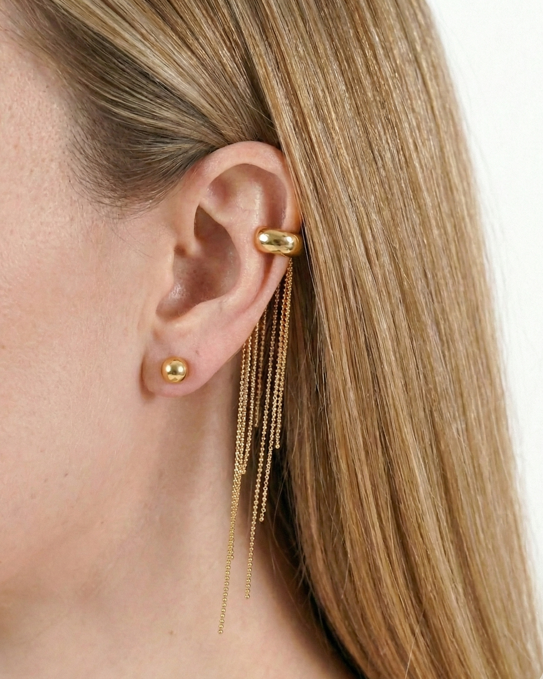 Gold earring with dangling chain on a person's ear against a neutral background