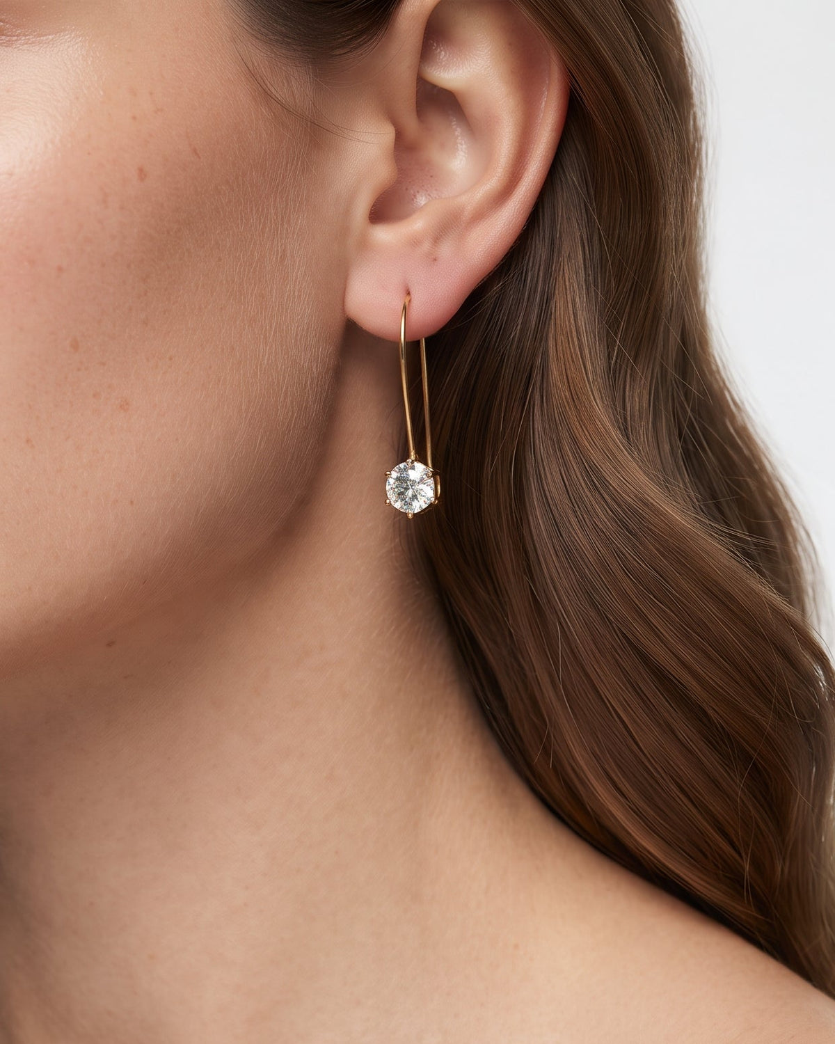 A close-up of a woman wearing a delicate gold dangle earring with a clear round gemstone, shown against a clean white background.