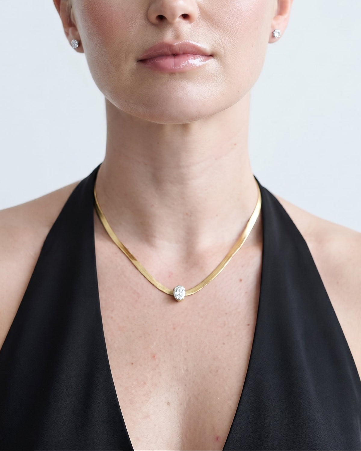 A blonde woman wearing a delicate gold chain necklace with a small clear gemstone pendant, styled with a black halter top against a light gray background.