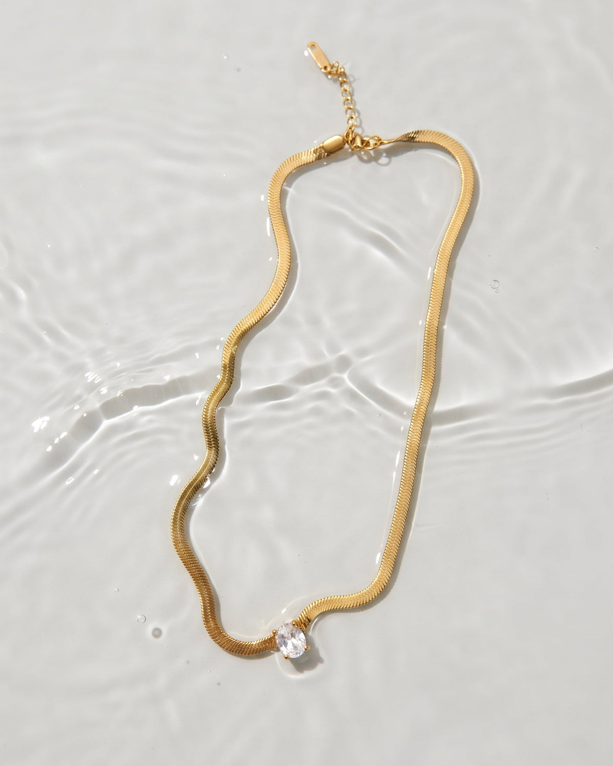 A gold snake chain necklace with a single clear faceted gemstone pendant, laid flat on a light gray surface with subtle water ripples.