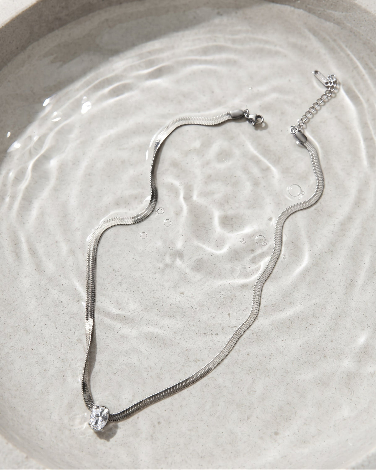 A silver snake chain necklace with a clear crystal pendant, displayed inside a shallow, textured gray ceramic bowl filled with water.