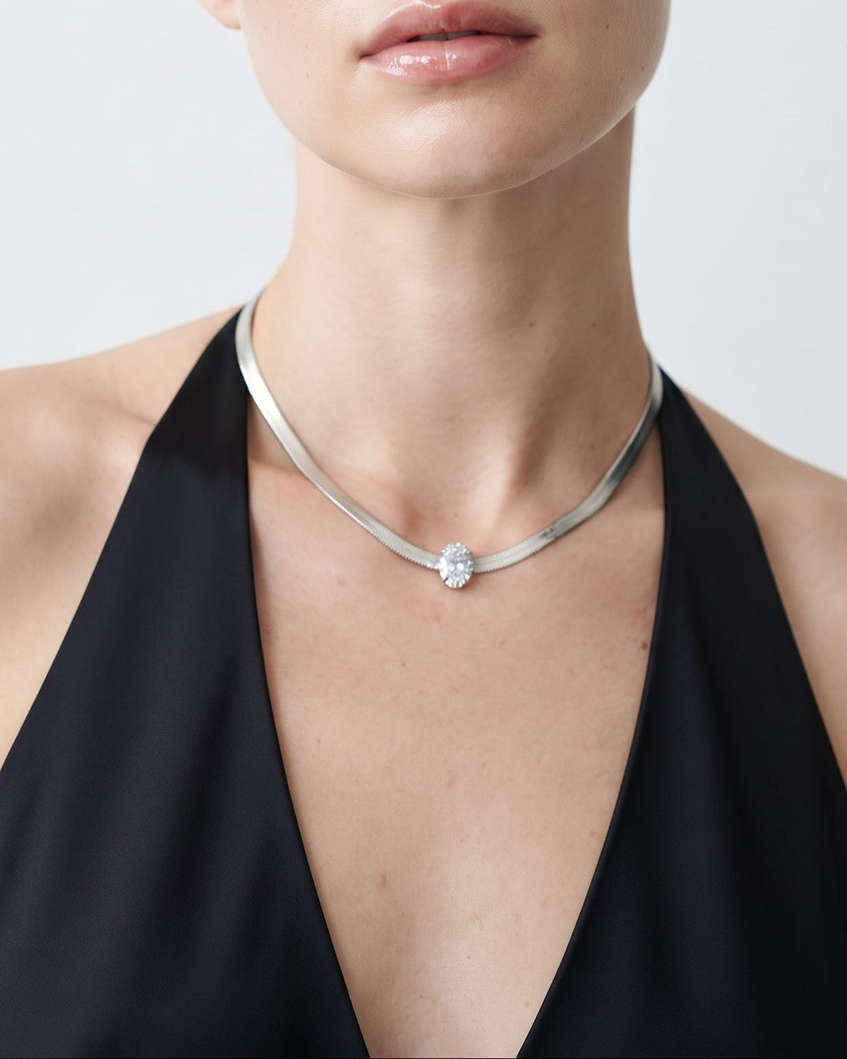 A blonde model wearing a thin silver chain necklace with a small clear gemstone pendant, paired with a black halter top against a light gray background.