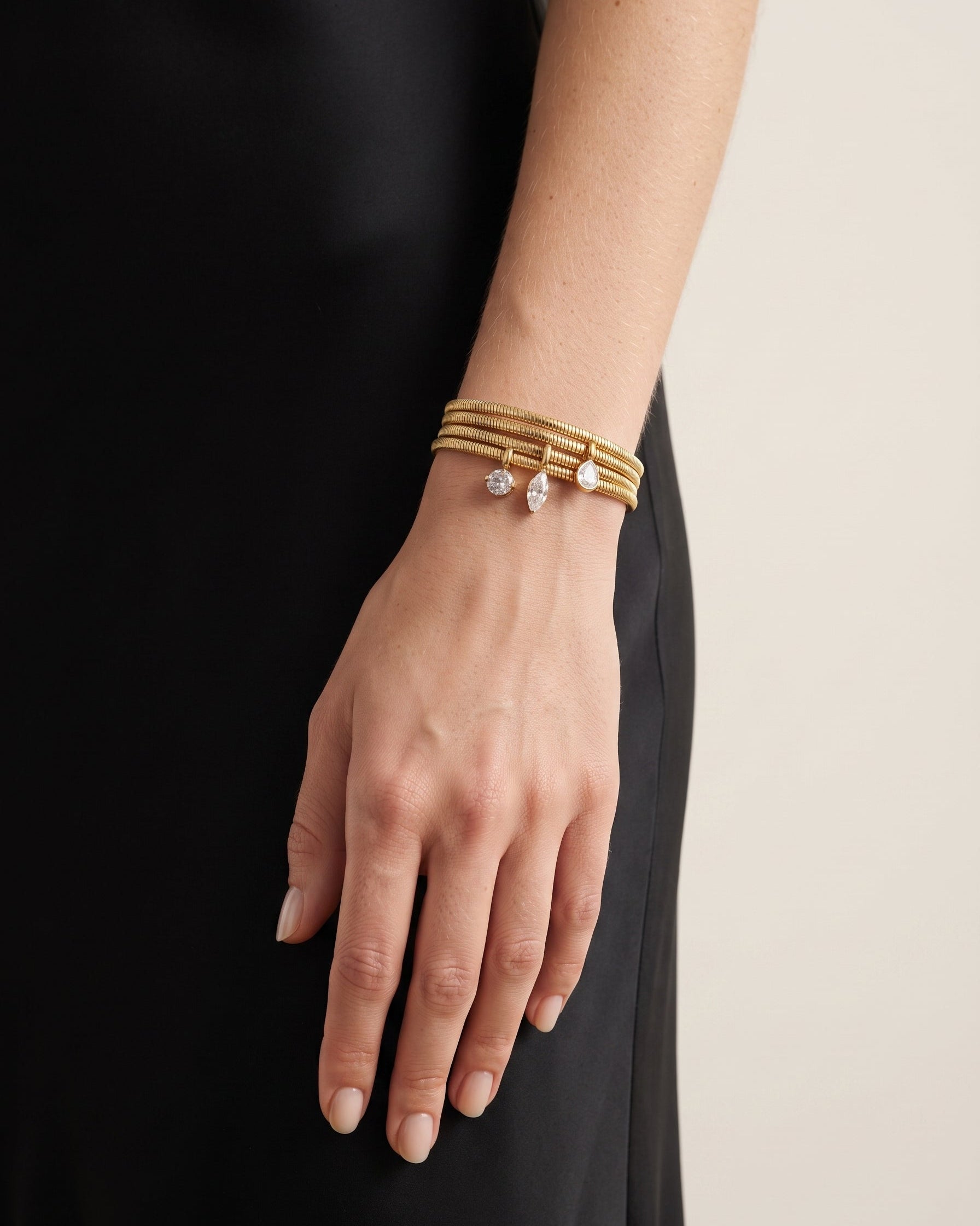 A close-up of a person's hand and wrist wearing a stack of gold bracelets, including one with a small clear crystal charm, against a black dress and a light beige background.