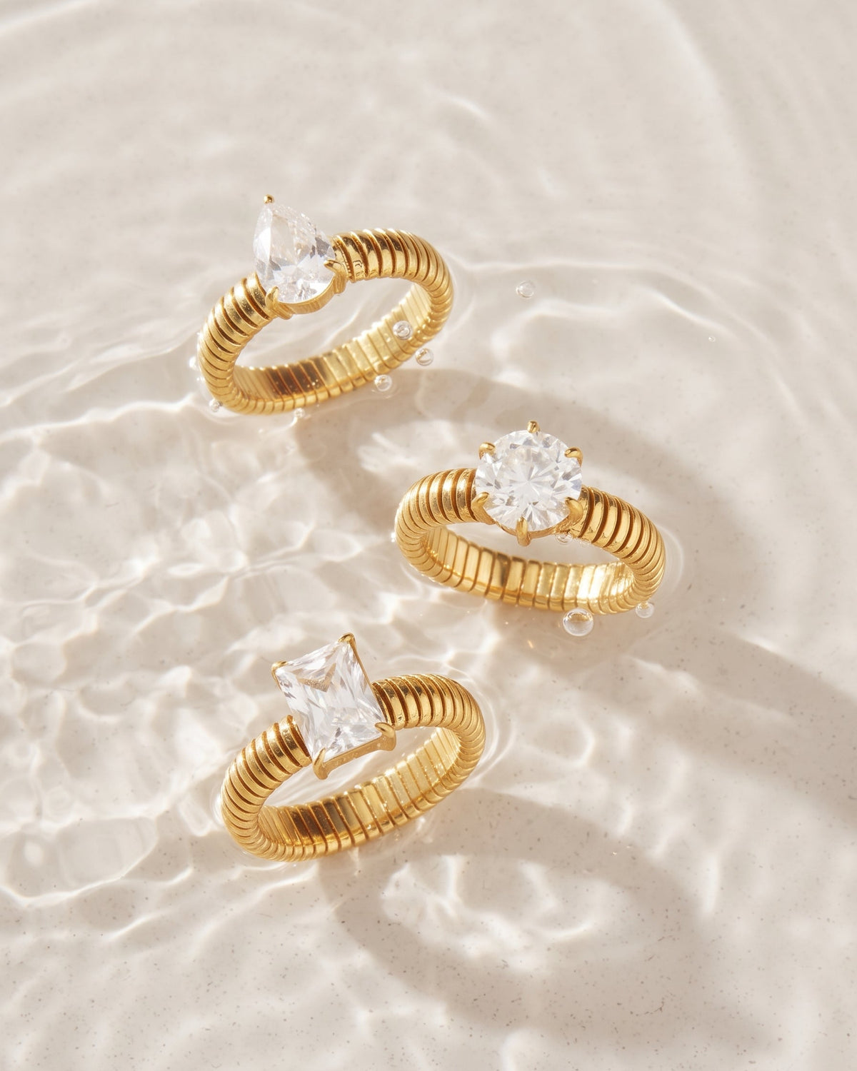 Three gold rings with clear gemstones resting on a light beige sandy surface with shallow water ripples. The rings feature textured bands and different stone shapes: marquise, round, and princess-cut.