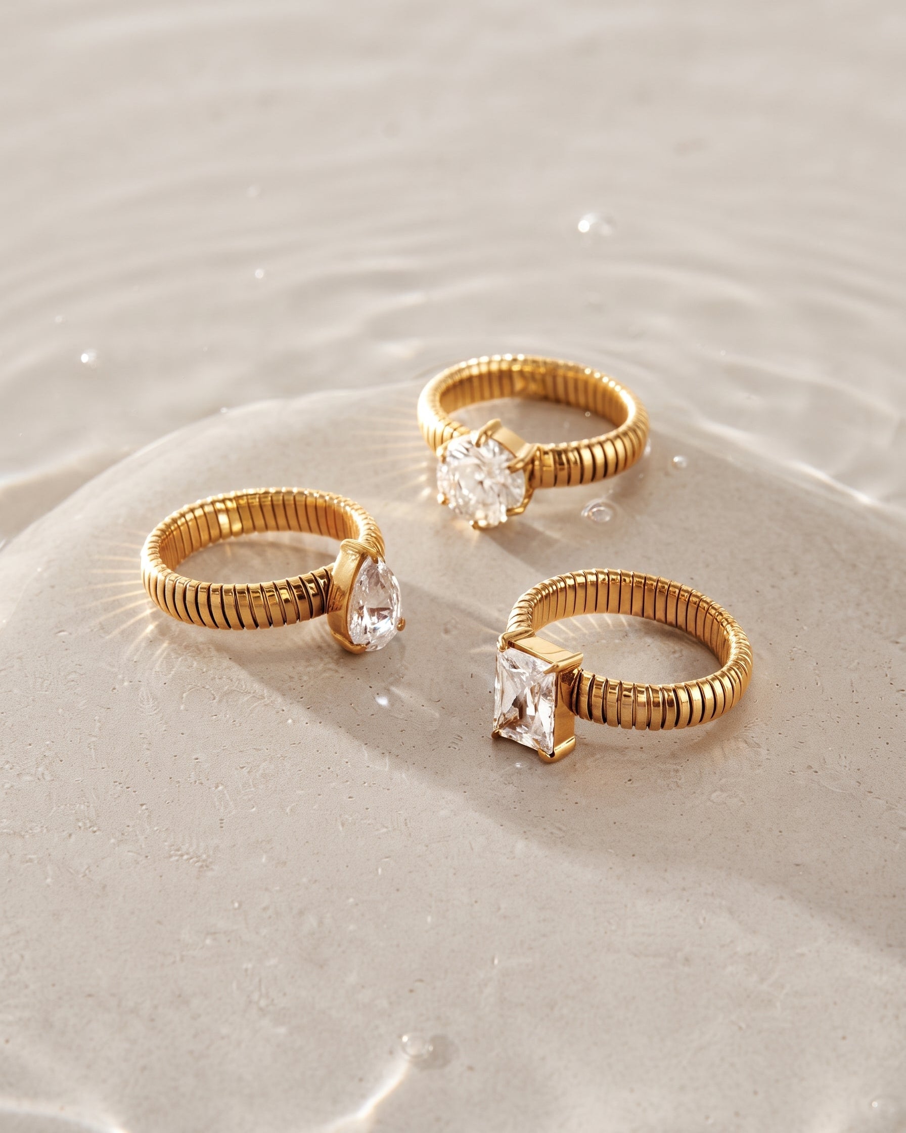 Three gold rings with clear gemstones arranged on wet sand with shallow water ripples. The rings feature textured bands and rectangular and teardrop-cut clear stones.