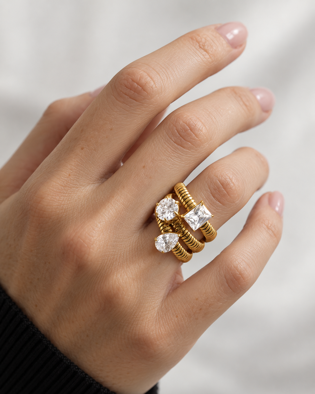 A close-up of a hand wearing a stack of three gold rings, including two rings with clear gemstones, against a soft white background.