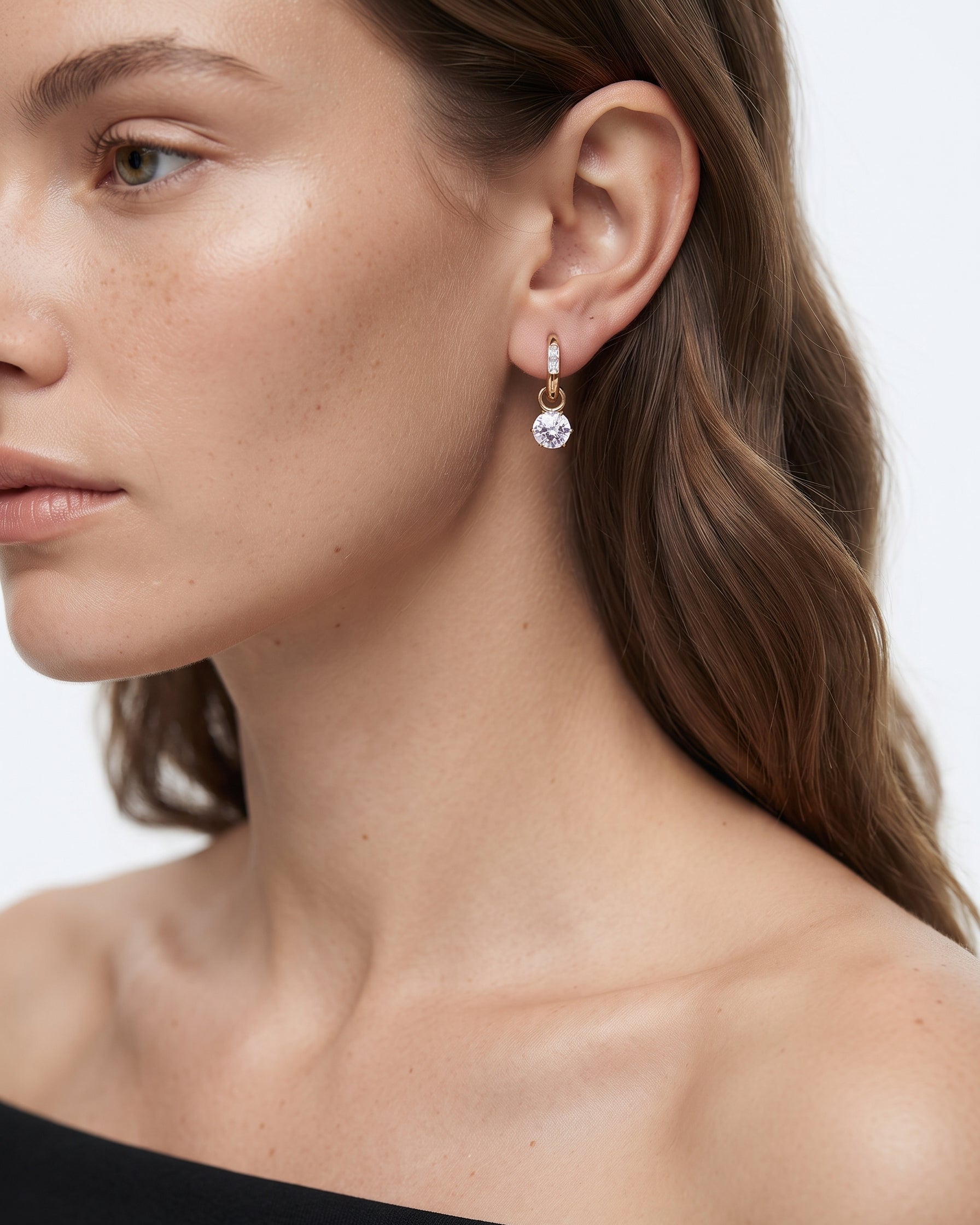 A close-up of a woman wearing a delicate gold huggie hoop earring with a dangling clear faceted gemstone, shown against a clean white background.