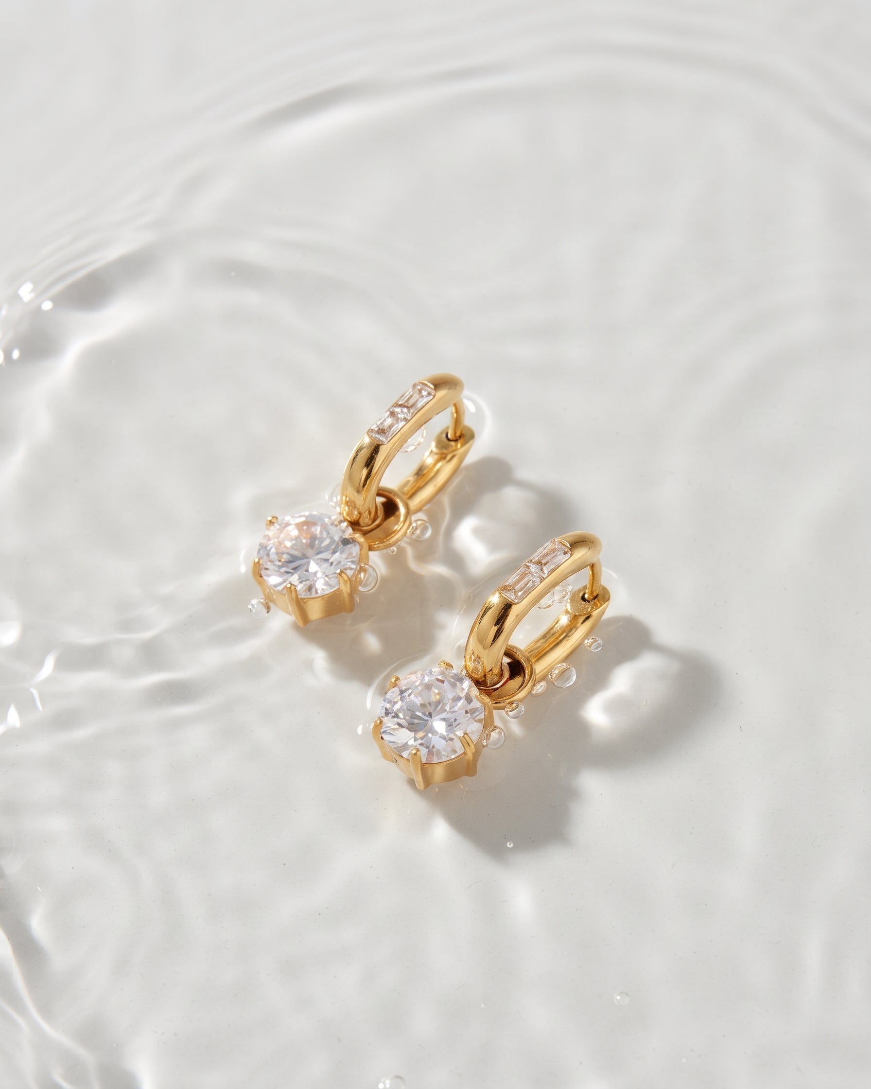 A pair of gold hoop earrings with large clear round-cut gemstones and small side stones, photographed on a glossy white surface with soft water ripples around them.