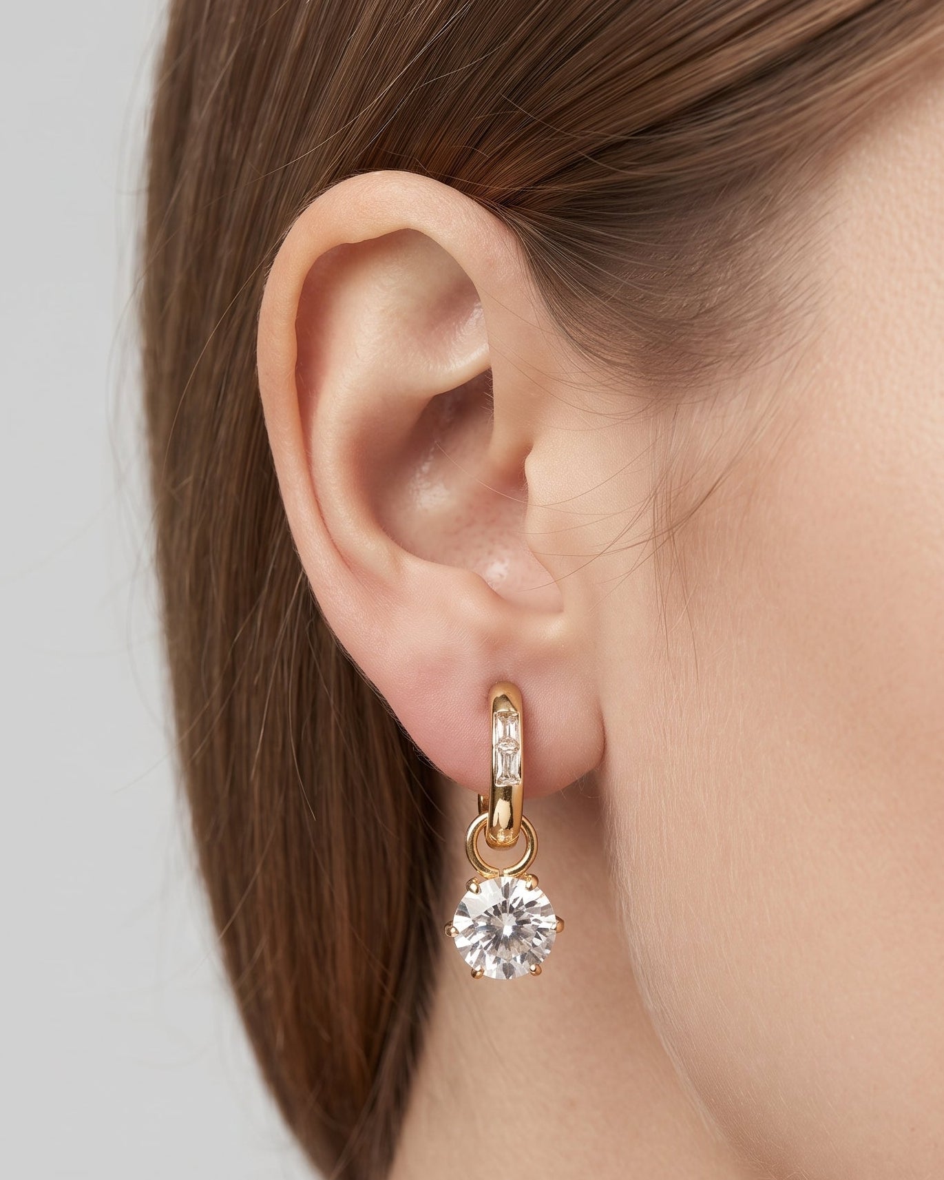 A close-up of a model's ear wearing a delicate gold huggie hoop earring with a small clear gemstone and a larger round clear gemstone dangle.