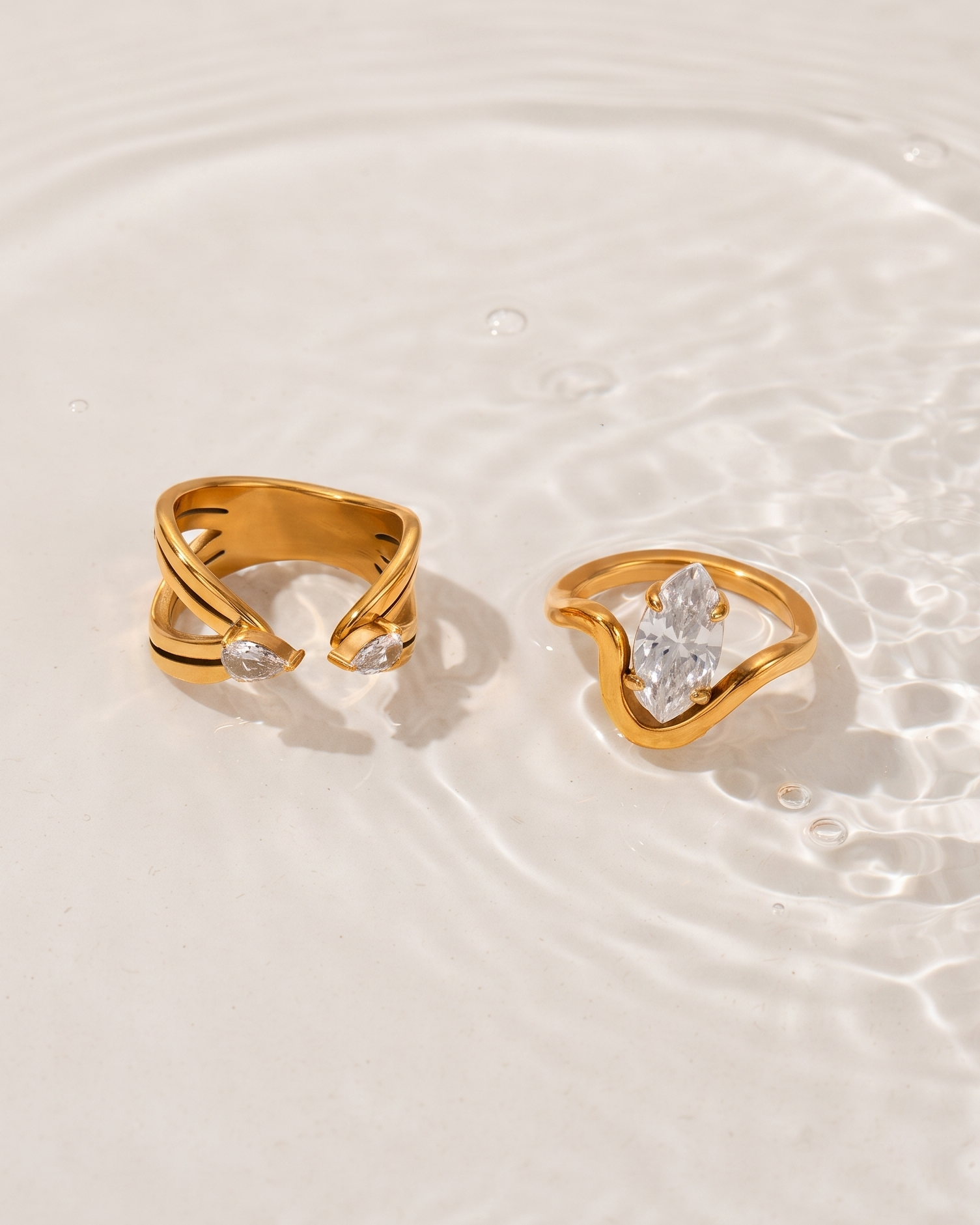 Two gold rings with clear gemstones, resting on a light beige surface with soft water ripples in the background.