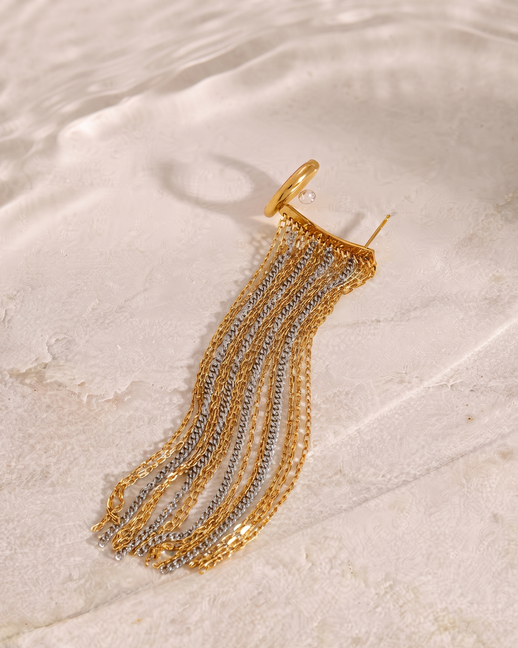A gold and silver chain fringe earring with a single gold hoop, displayed on a textured white stone surface.