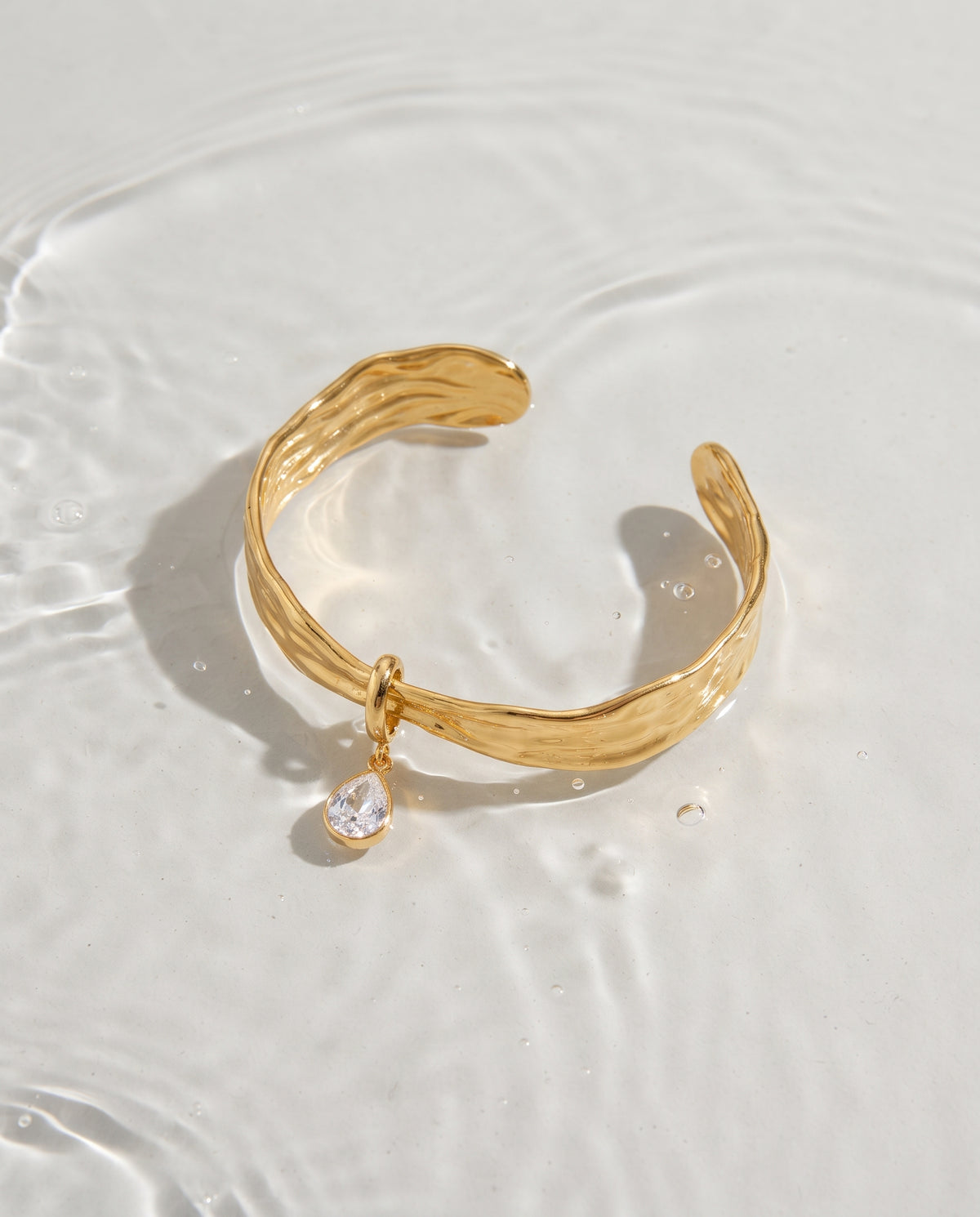 A gold-toned open cuff bracelet with a textured, hammered finish and a small teardrop-shaped clear crystal charm, resting on a white surface with water ripples.