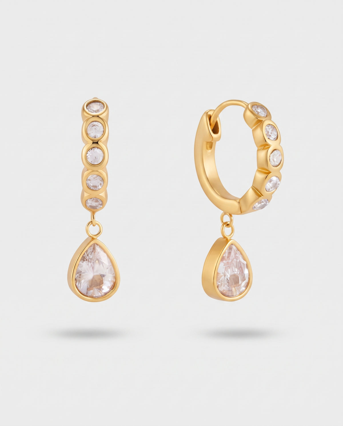 A pair of gold huggie hoop earrings with clear gemstone accents and teardrop-shaped clear crystal drops, shown on a light gray background.