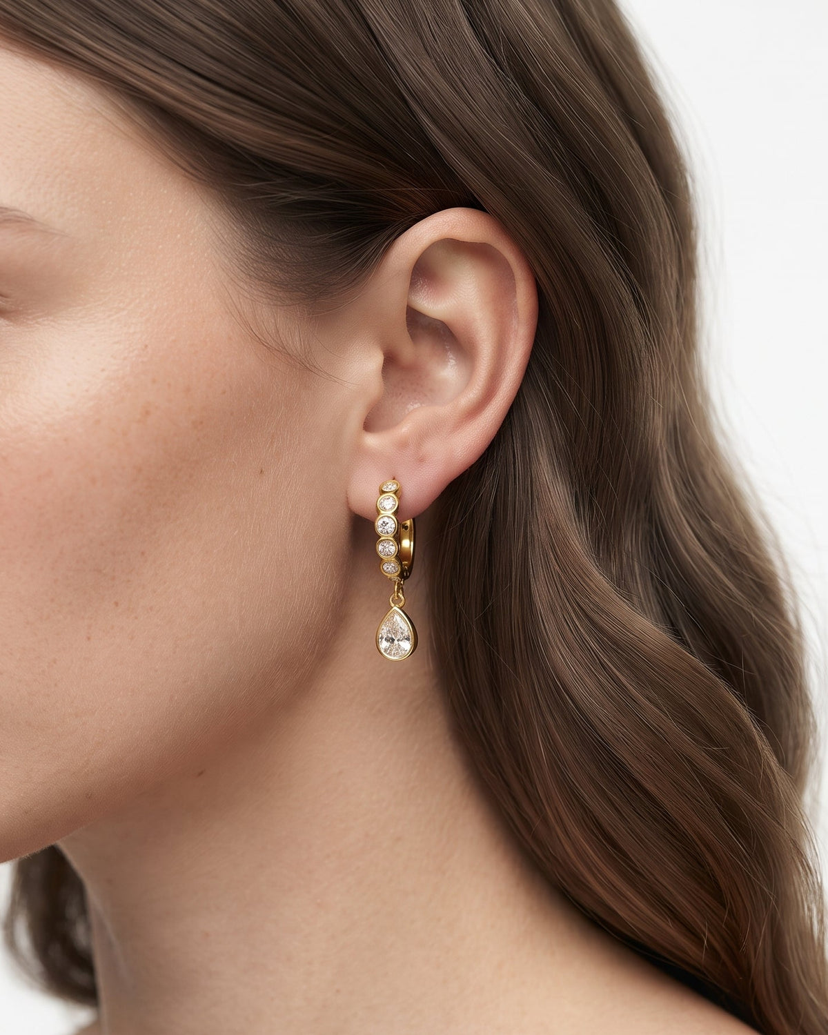A close-up side profile of a woman wearing a delicate gold hoop earring adorned with small clear stones and a teardrop-shaped clear drop, against a clean white background.
