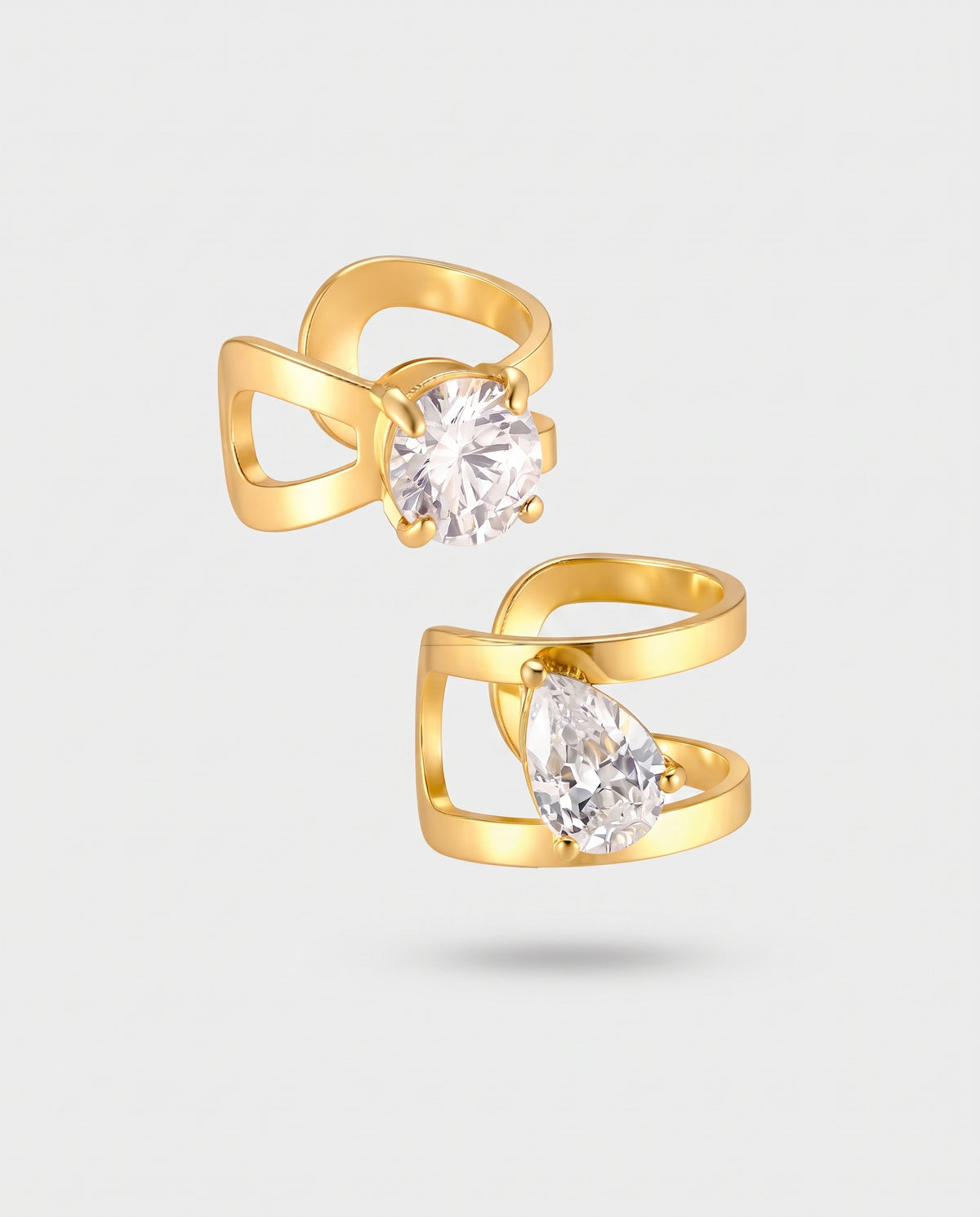 A pair of gold-plated ring earrings with open wrap-around bands and clear gemstones, one round and one pear-shaped, set against a light gray background.