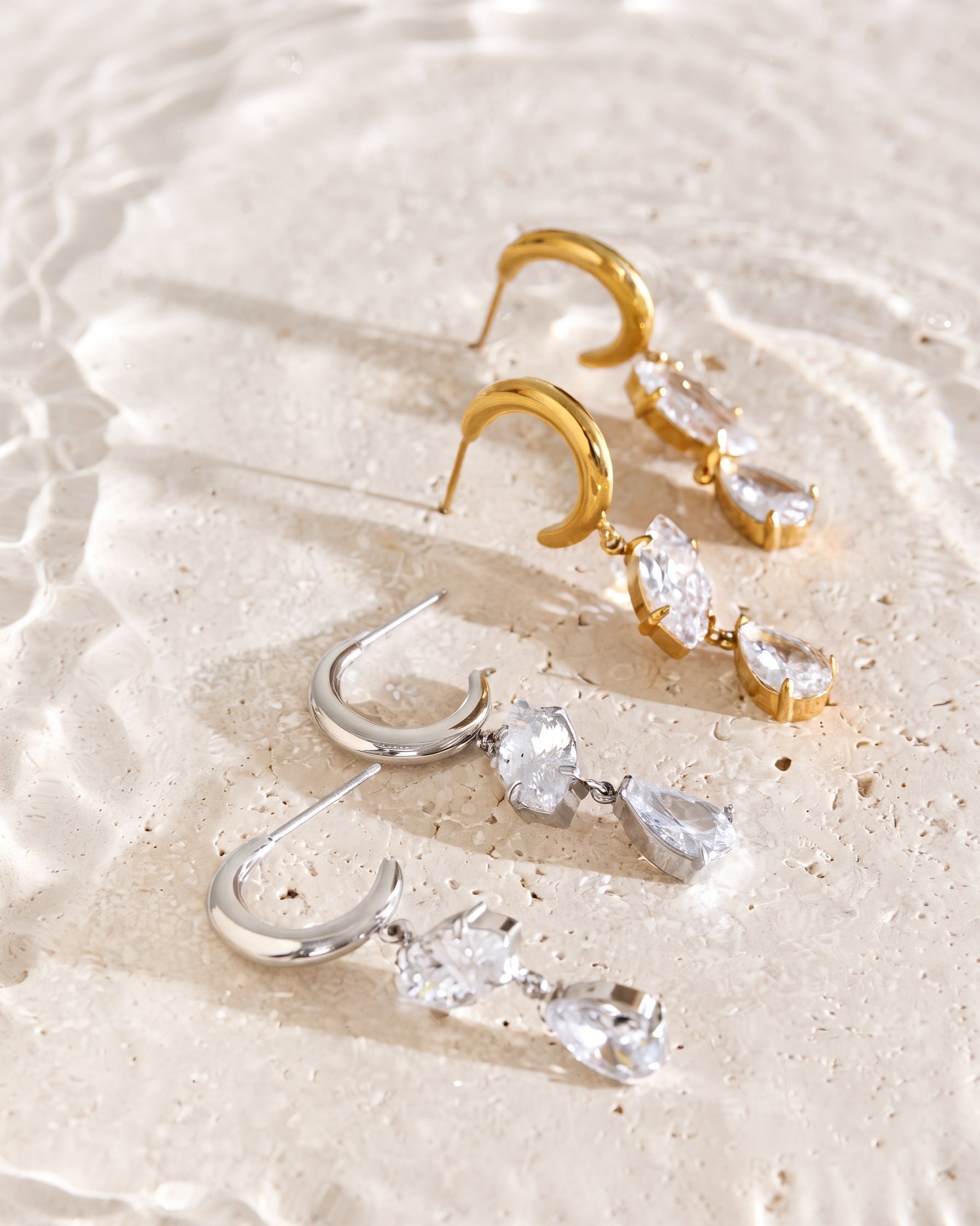 A pair of gold crescent drop earrings with clear teardrop crystals and a pair of silver crescent drop earrings with clear teardrop crystals, displayed on a textured beige surface with a shallow depth of field.