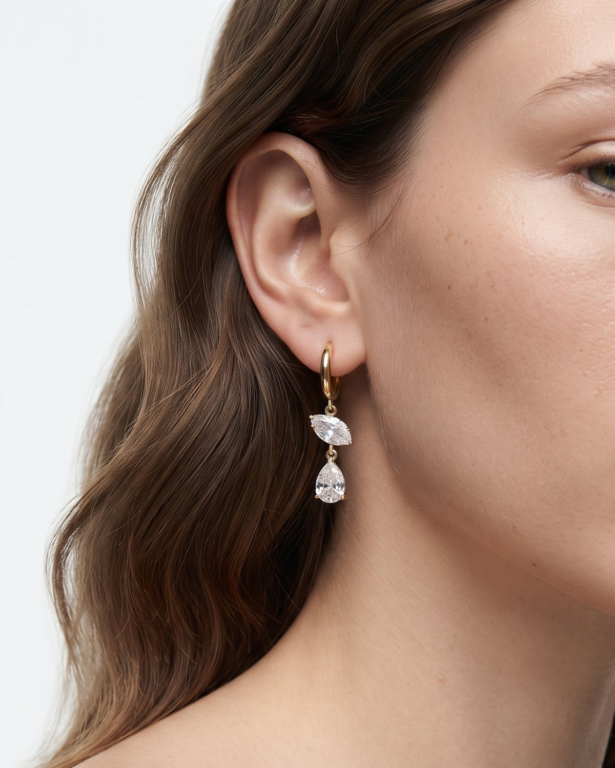 A close-up of a woman wearing a delicate gold huggie hoop earring with two cascading pear-cut clear gemstones, set against a clean white background.
