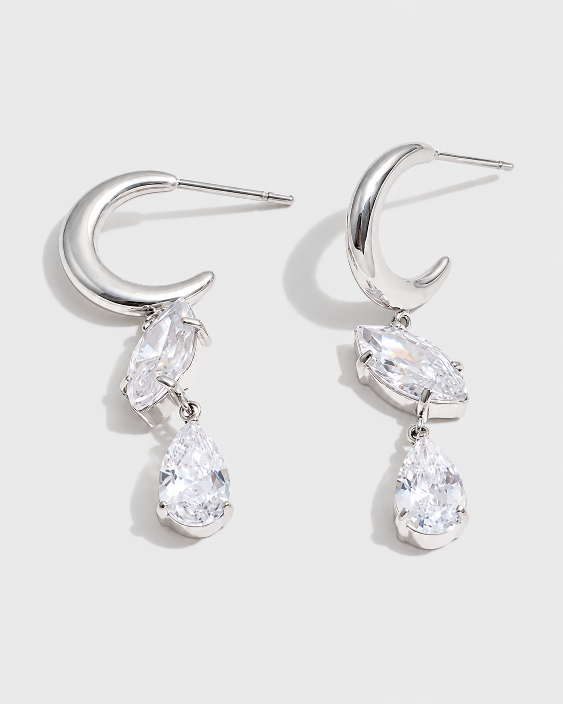 A pair of silver crescent-shaped drop earrings with marquise and pear-cut clear gemstones, displayed on a clean light gray background.