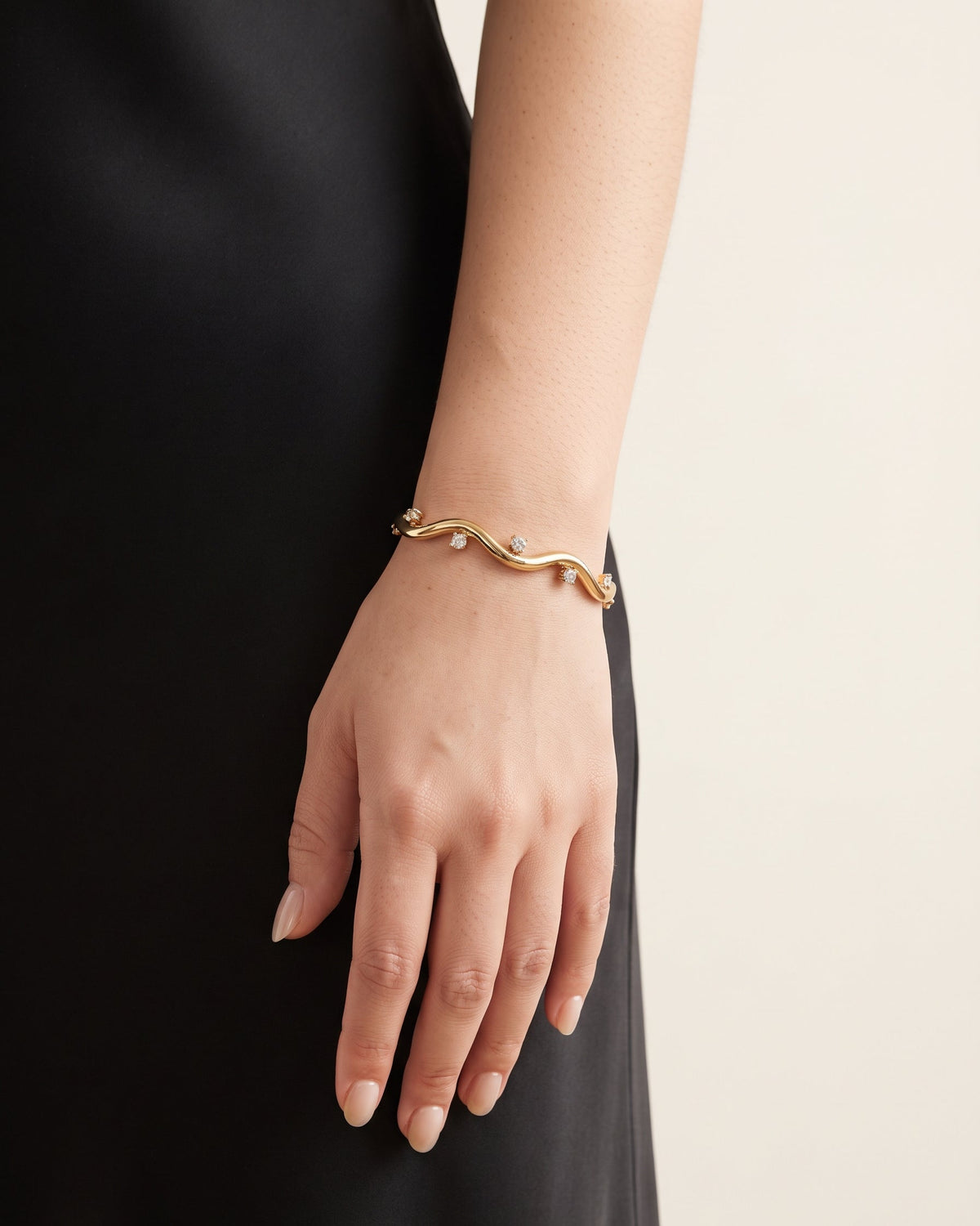 A close-up of a person's hand and wrist wearing a delicate gold wavy bracelet with small clear stones, set against a black dress and a light beige background.