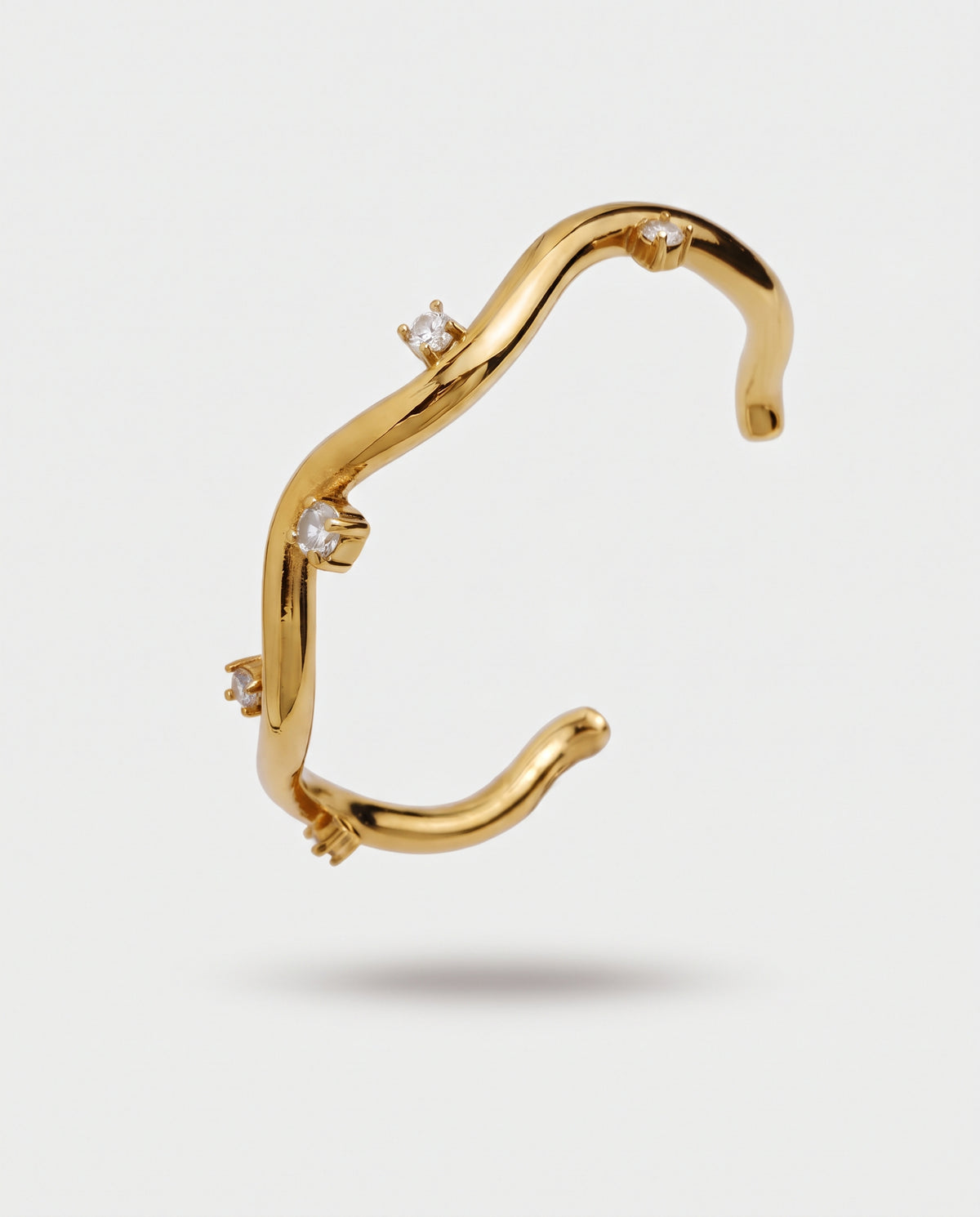 A gold open cuff bracelet with a wavy, organic curve and four small clear gemstones set along the front, shown against a light gray background.