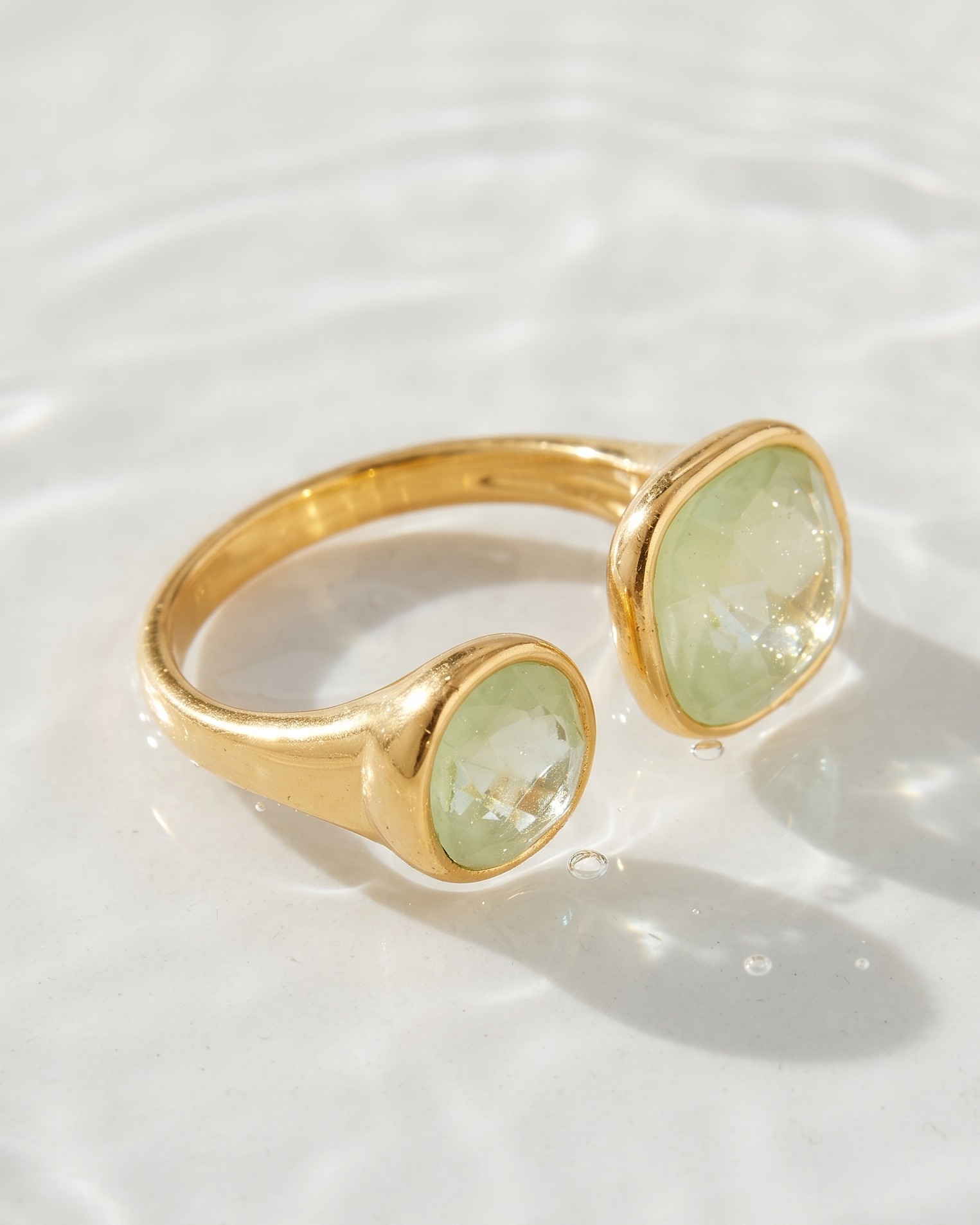 A gold ring with two oval light green gemstones, resting on a white surface with soft water ripples and shadows.