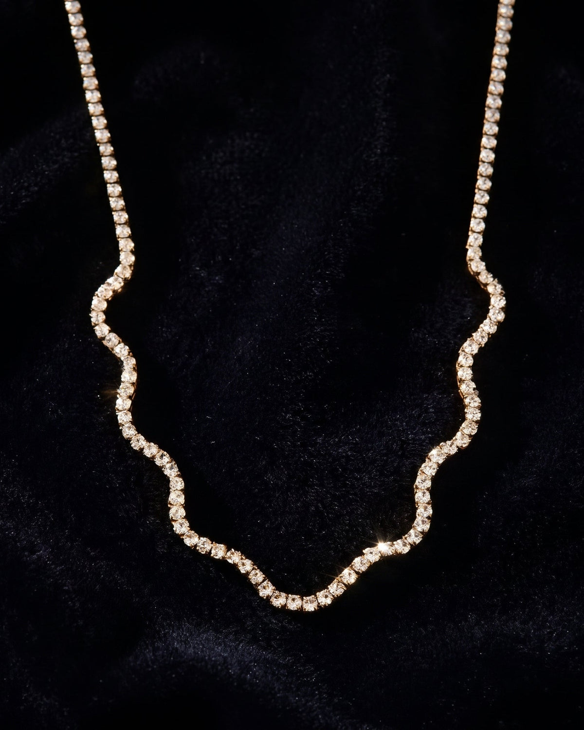 A delicate wavy chain necklace adorned with small clear rhinestones, displayed on a textured black velvet background.