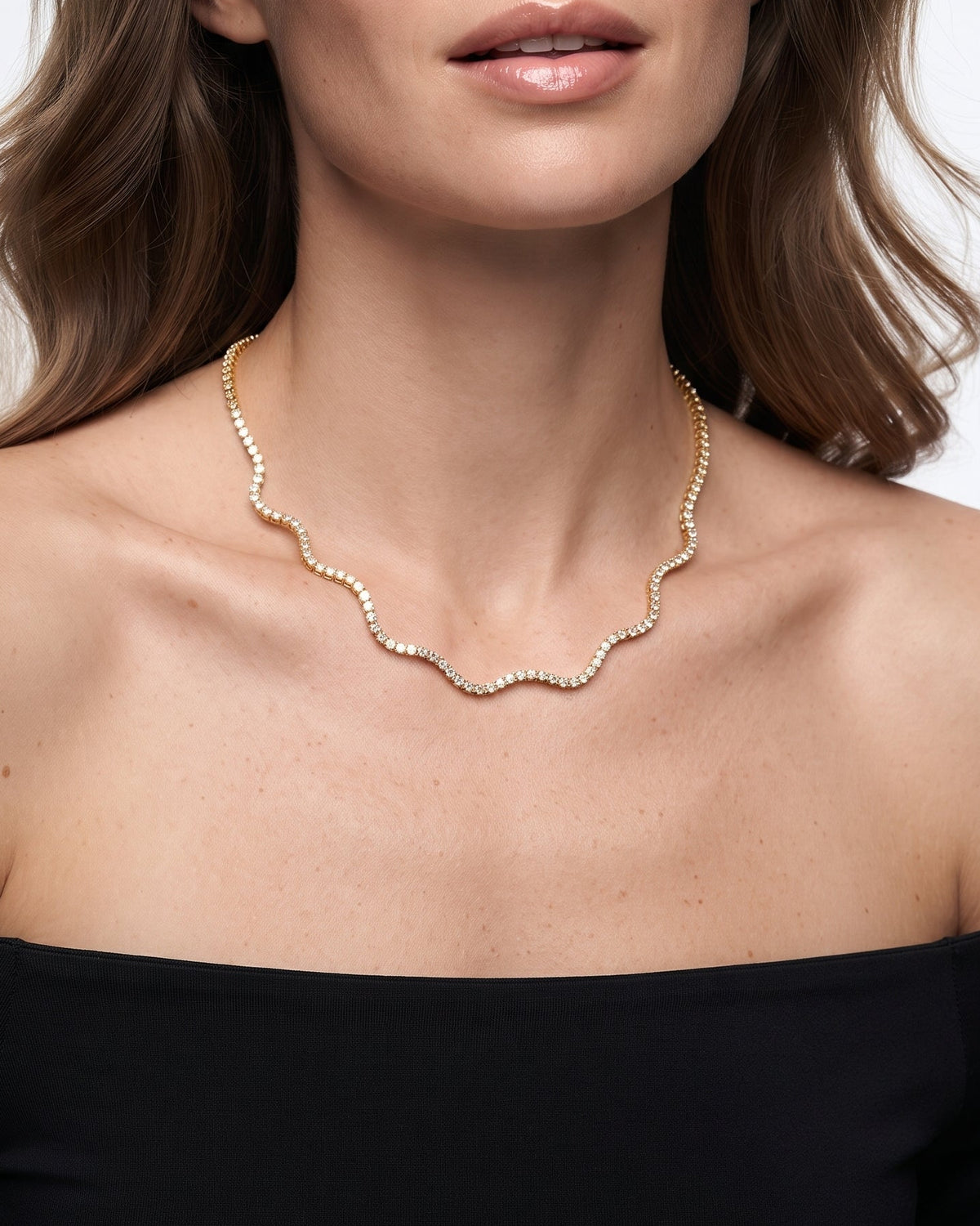 A woman wearing a delicate gold wavy chain necklace with small clear stones, styled with a black off-the-shoulder top against a light gray background.