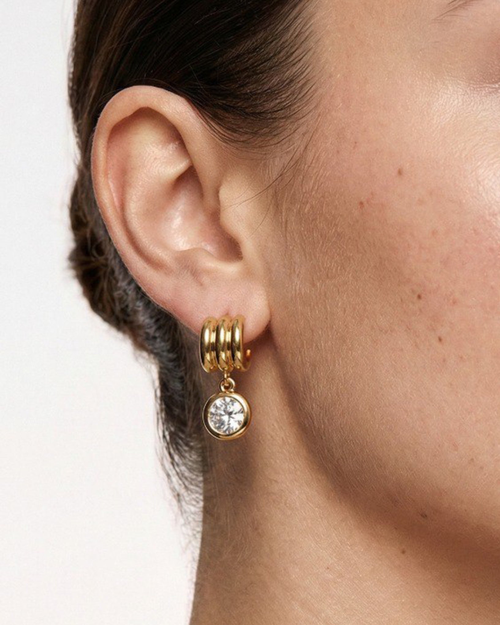 Gold hoop earring with a clear gemstone worn by a person.