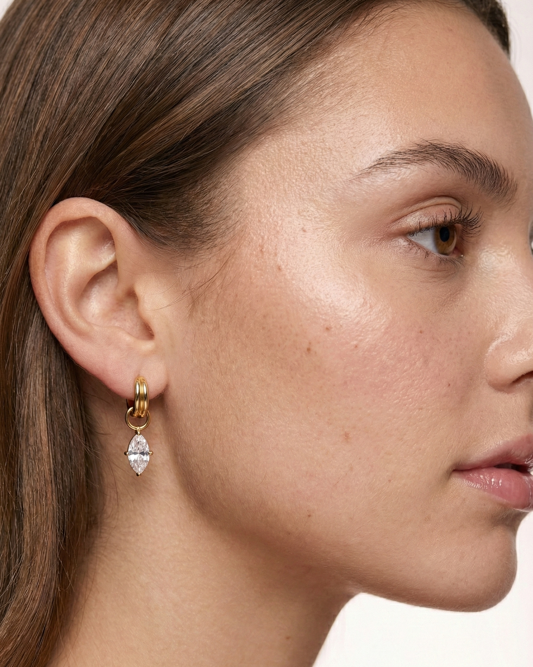 Close-up of a woman wearing gold earrings with a clear gemstone on a neutral background