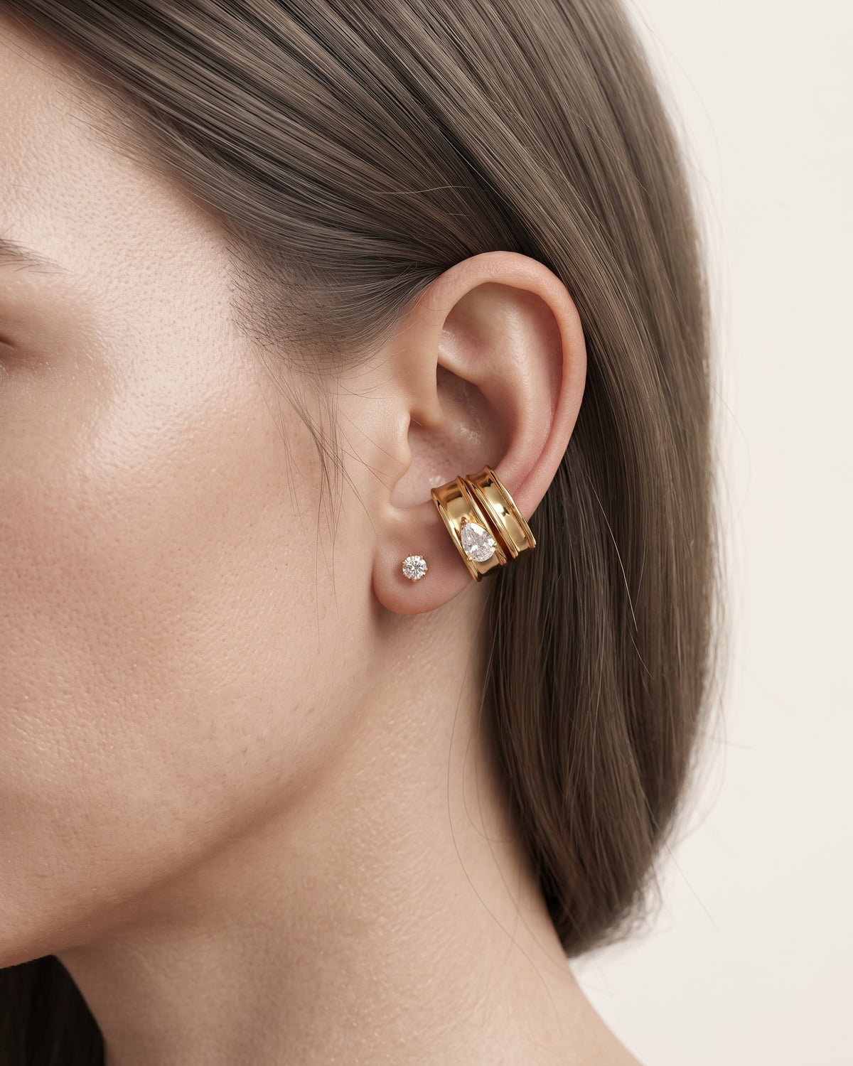 A close-up of a model's ear wearing a pair of gold huggie hoop earrings with clear pavé stones, alongside a small round diamond stud earring, against a soft beige background.