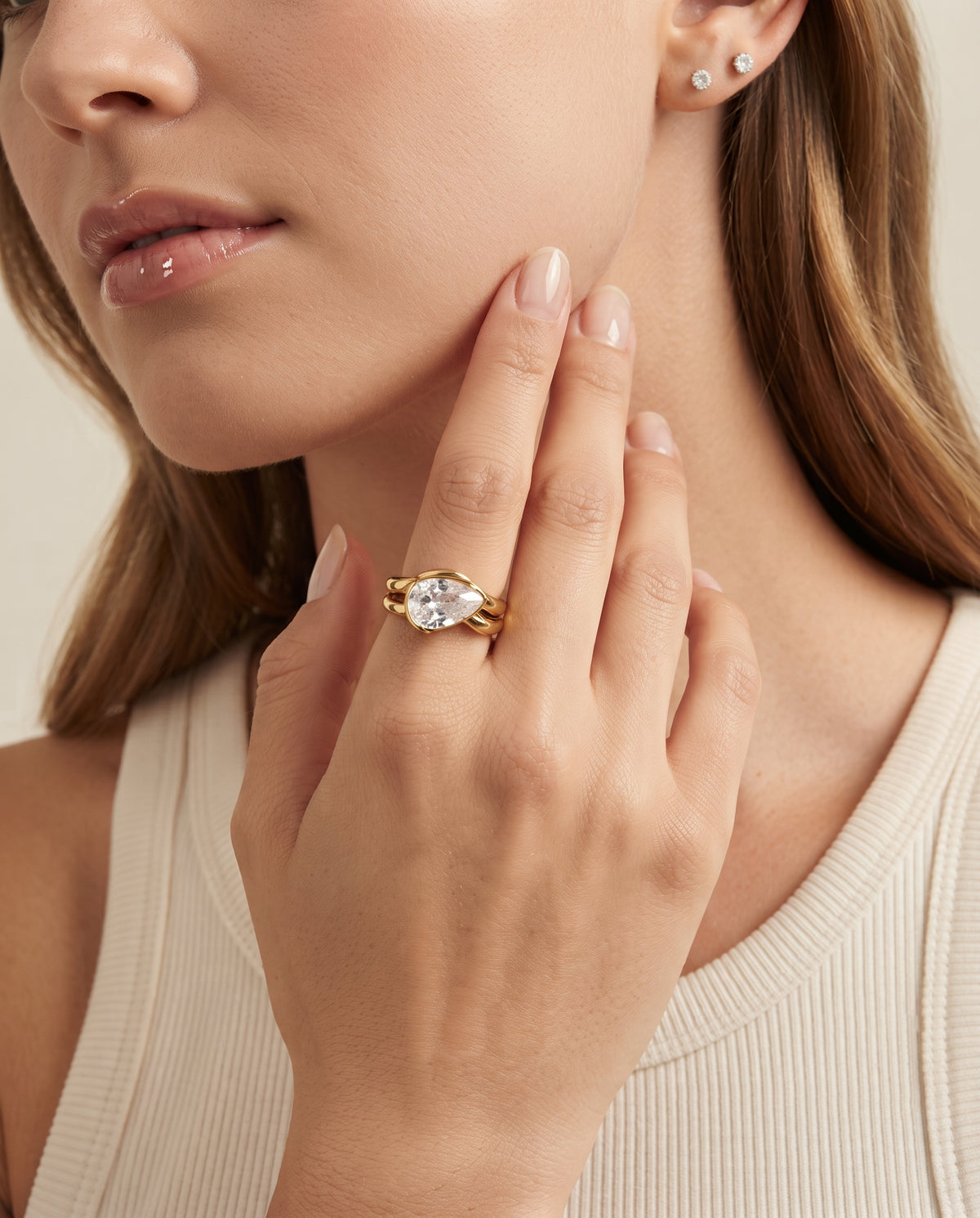 A close-up lifestyle shot of a model wearing a gold ring with a marquise-cut clear gemstone on her finger, paired with small stud earrings and a cream ribbed top.