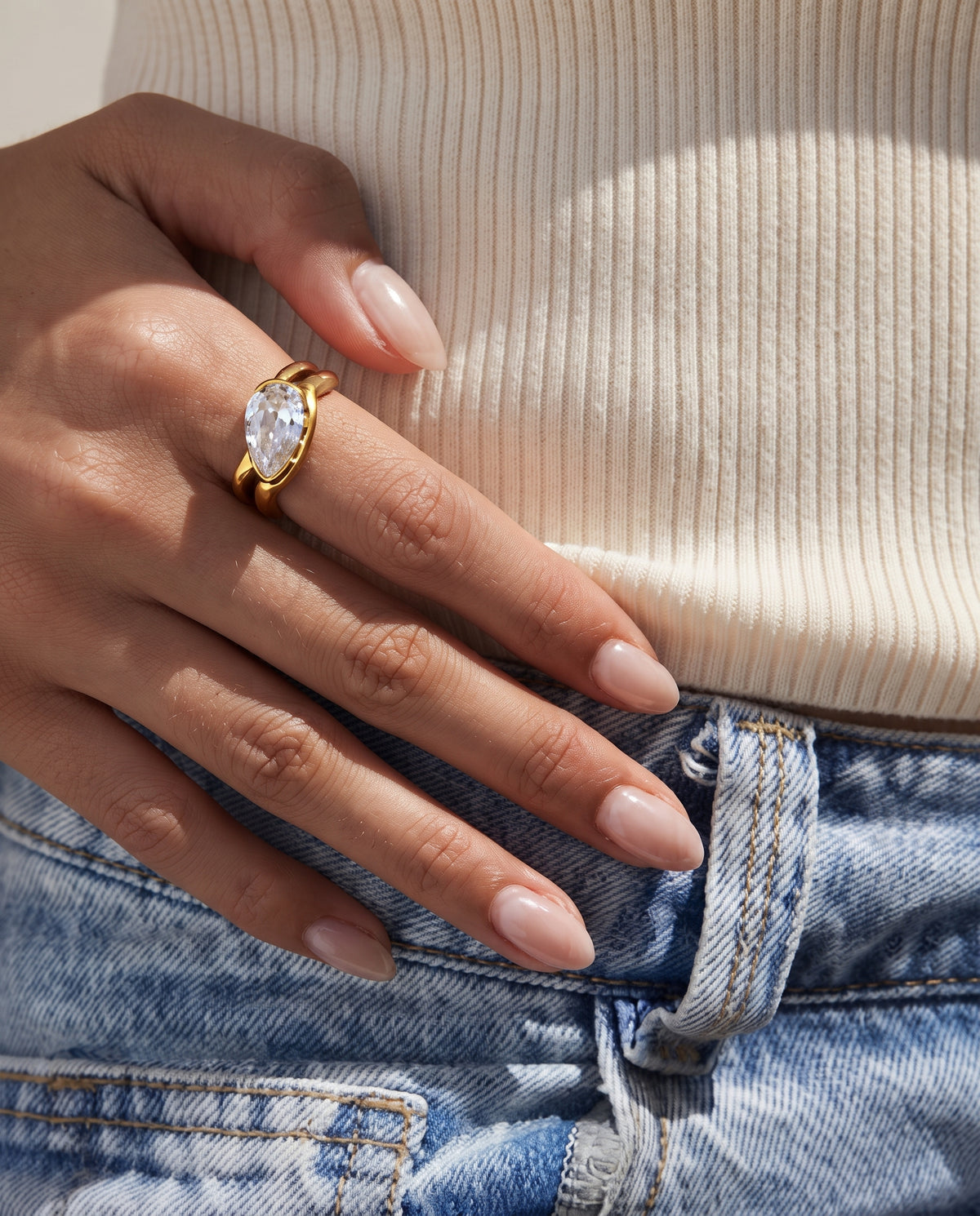 A close-up of a person's hand wearing a gold ring with a marquise-cut clear gemstone, styled with a cream ribbed top and blue denim jeans.